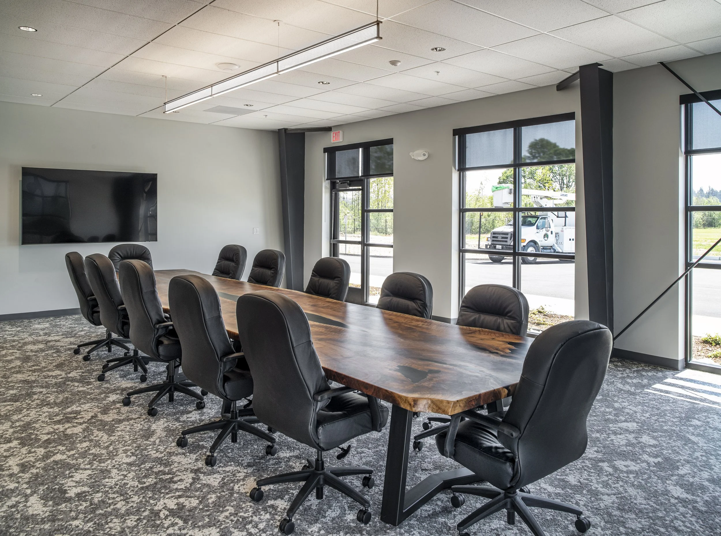 Modern conference room with a long wooden table, black leather chairs, wall-mounted TV, large windows with shades, and a view outside of a truck and greenery.