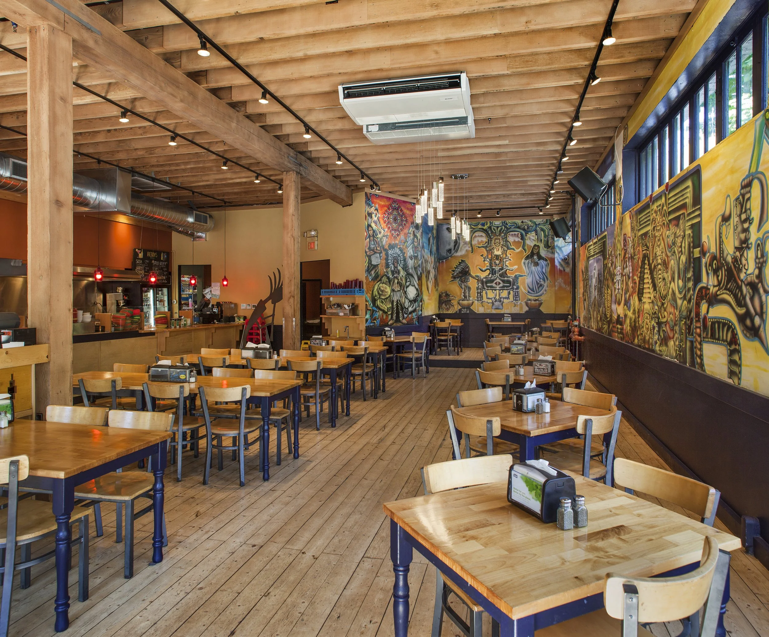 Interior of a restaurant with wooden tables and chairs, colorful murals on the walls, and warm lighting.