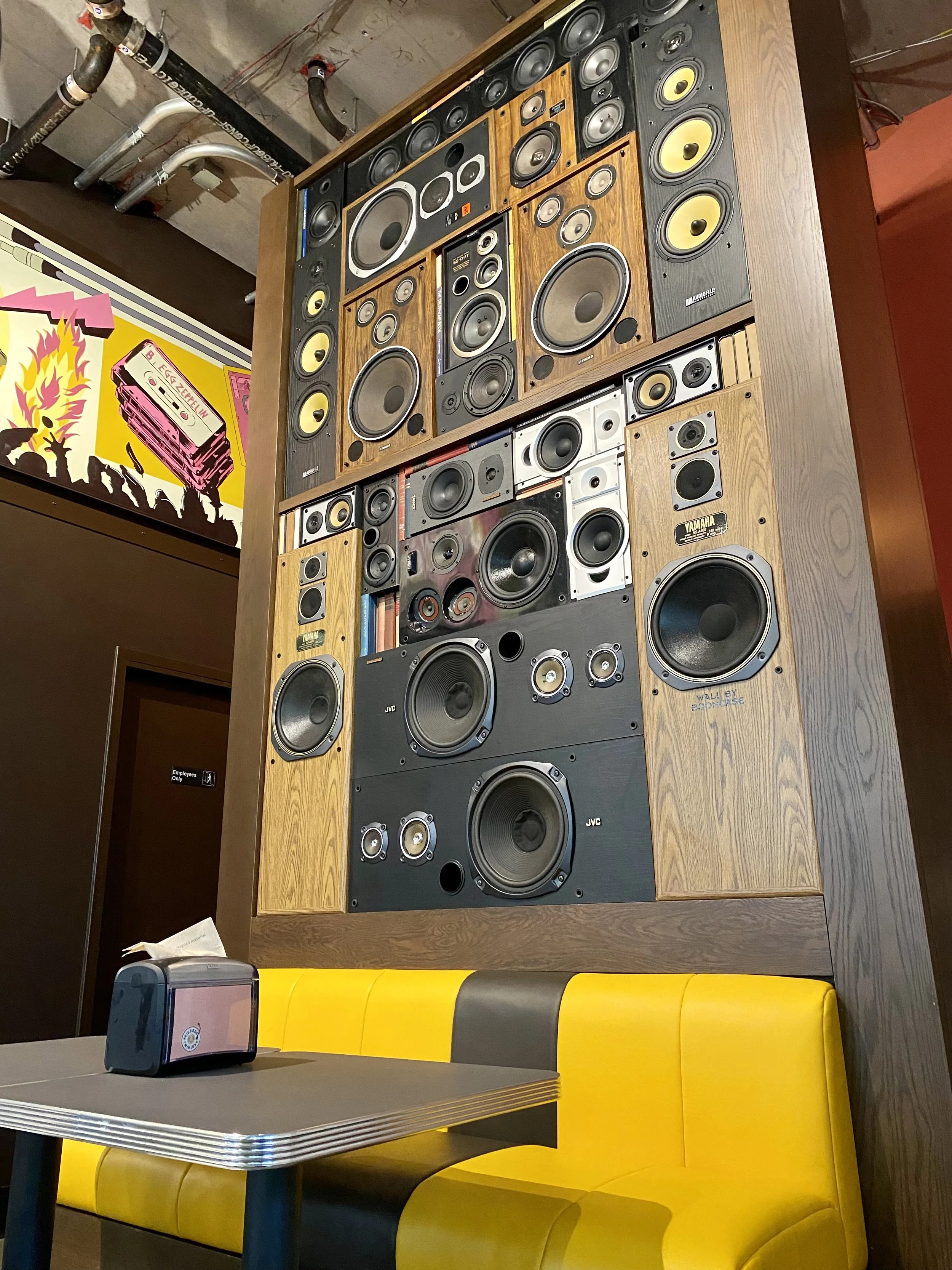 Large vintage wall of speaker systems in a room with yellow booth seating and a small table with a tissue box.
