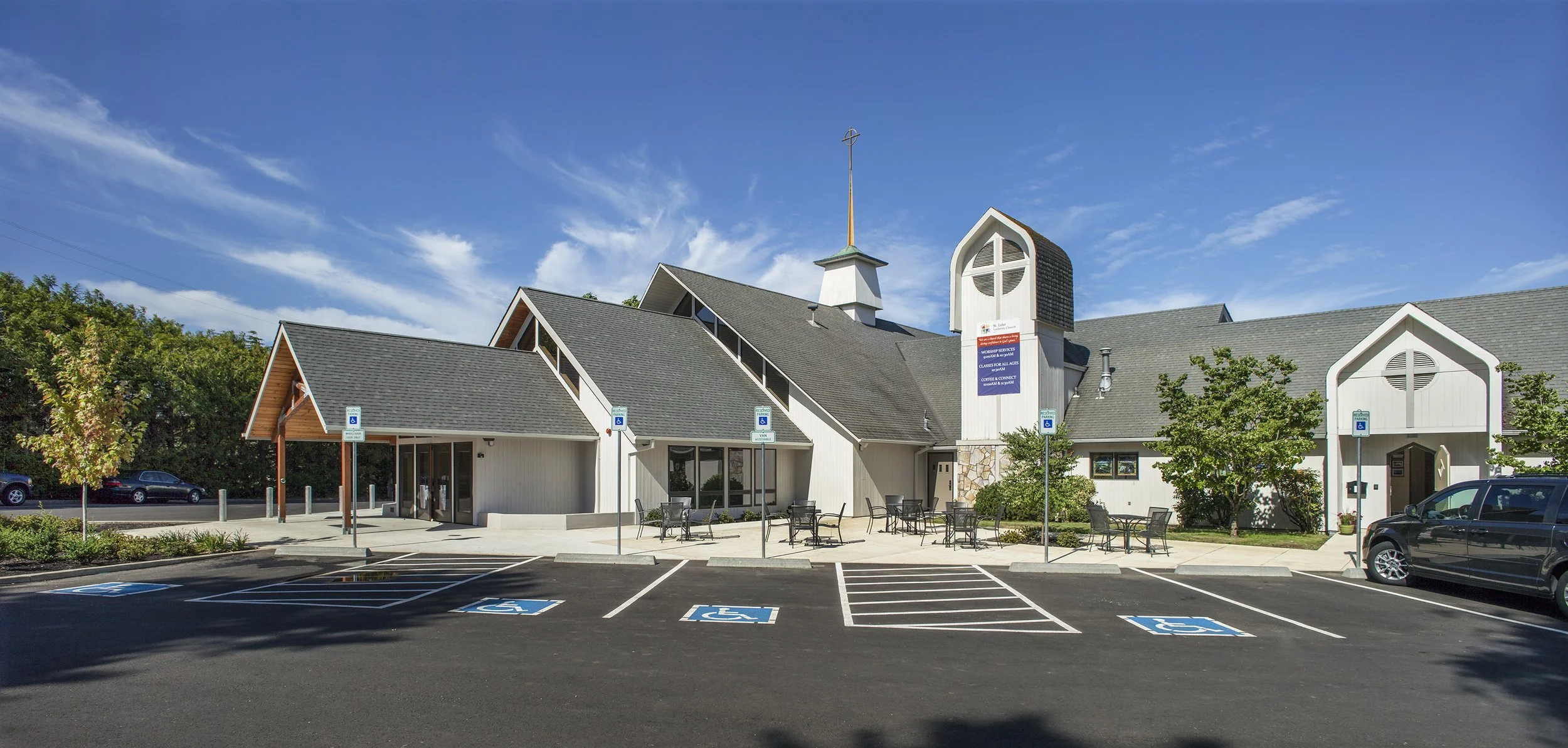 A church building with an adjacent parking lot, several accessible parking spaces, outdoor seating, trees, and a blue sky with clouds.