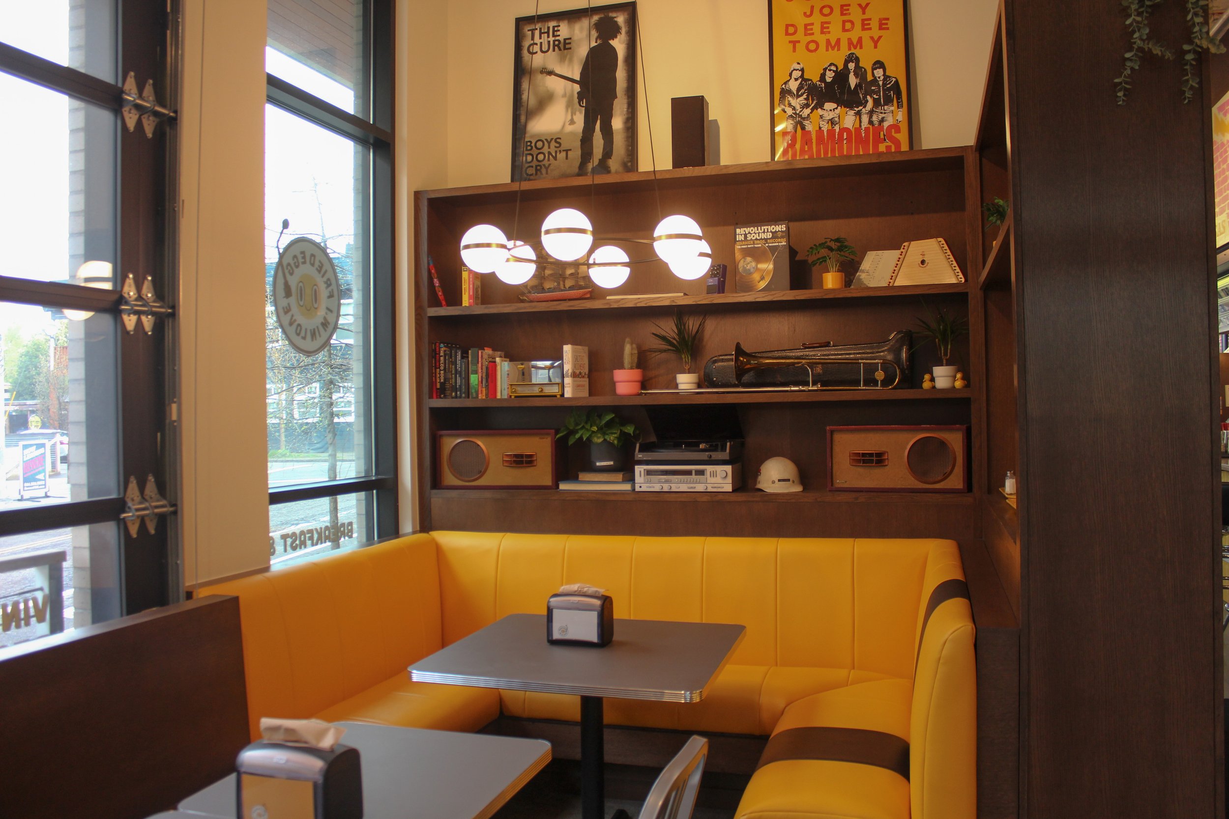 Interior of a diner with a yellow booth, black tables, a large window, and a wooden shelf filled with books, plants, a saxophone, and decorative items. Posters of The Cure, Joey Dee Dee, Tomy, and Ramones are on the wall above the shelf, with a moder