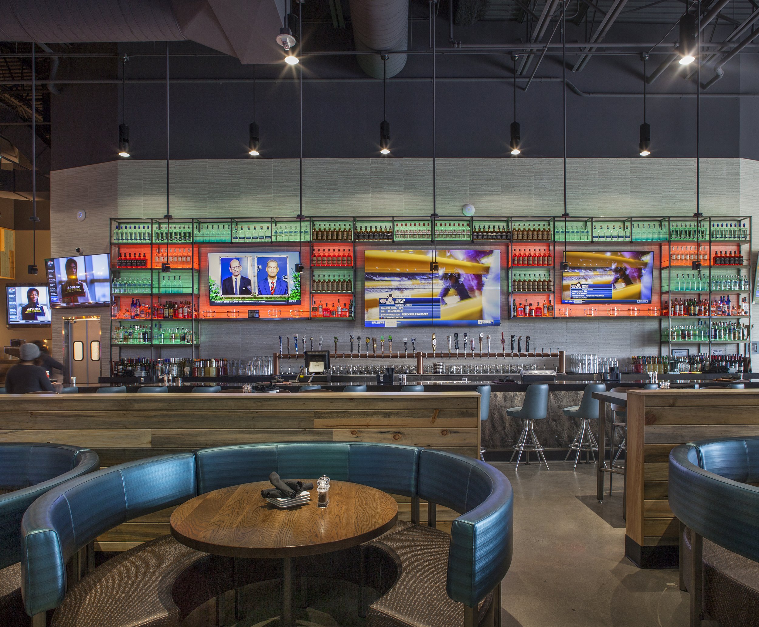Interior of a modern bar with multiple TV screens, a well-stocked bar counter, and seating areas with circular booths and chairs.