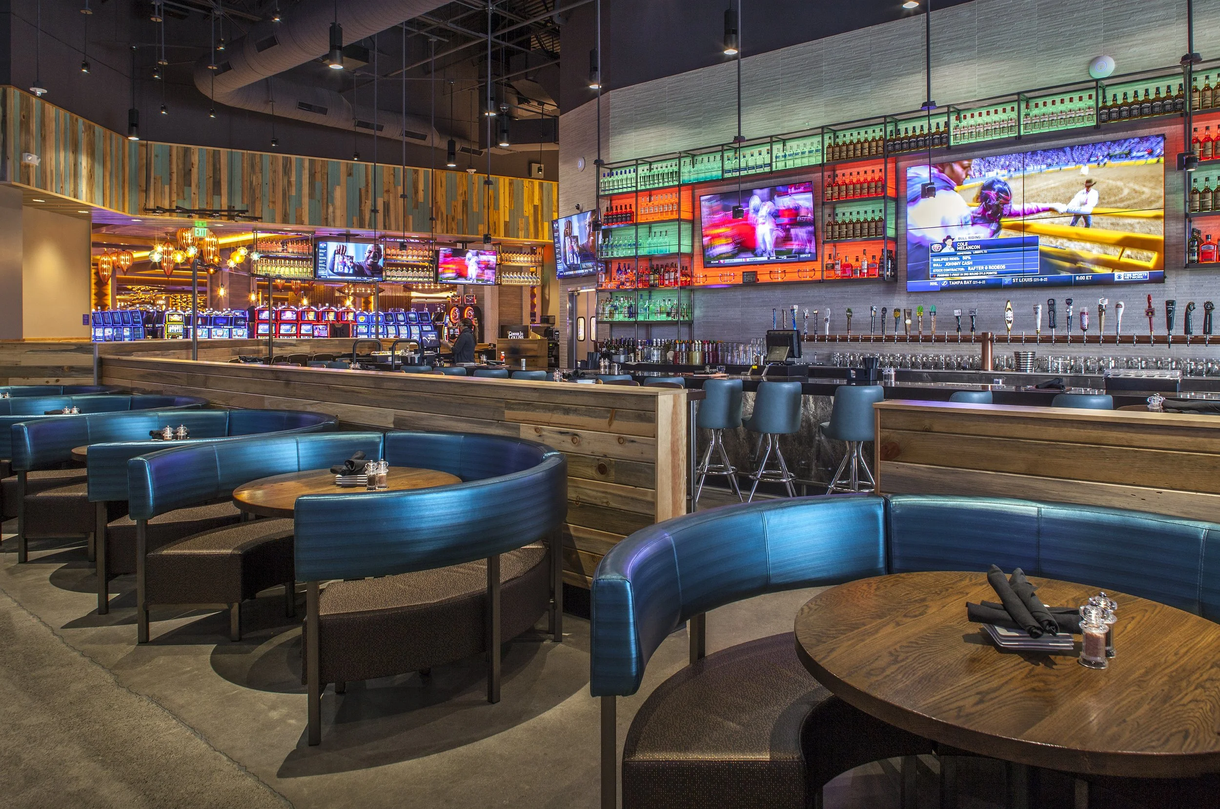 A modern bar and restaurant with blue booths, wooden tables, multiple TV screens showing sports, and a colorful array of alcohol bottles behind the bar.