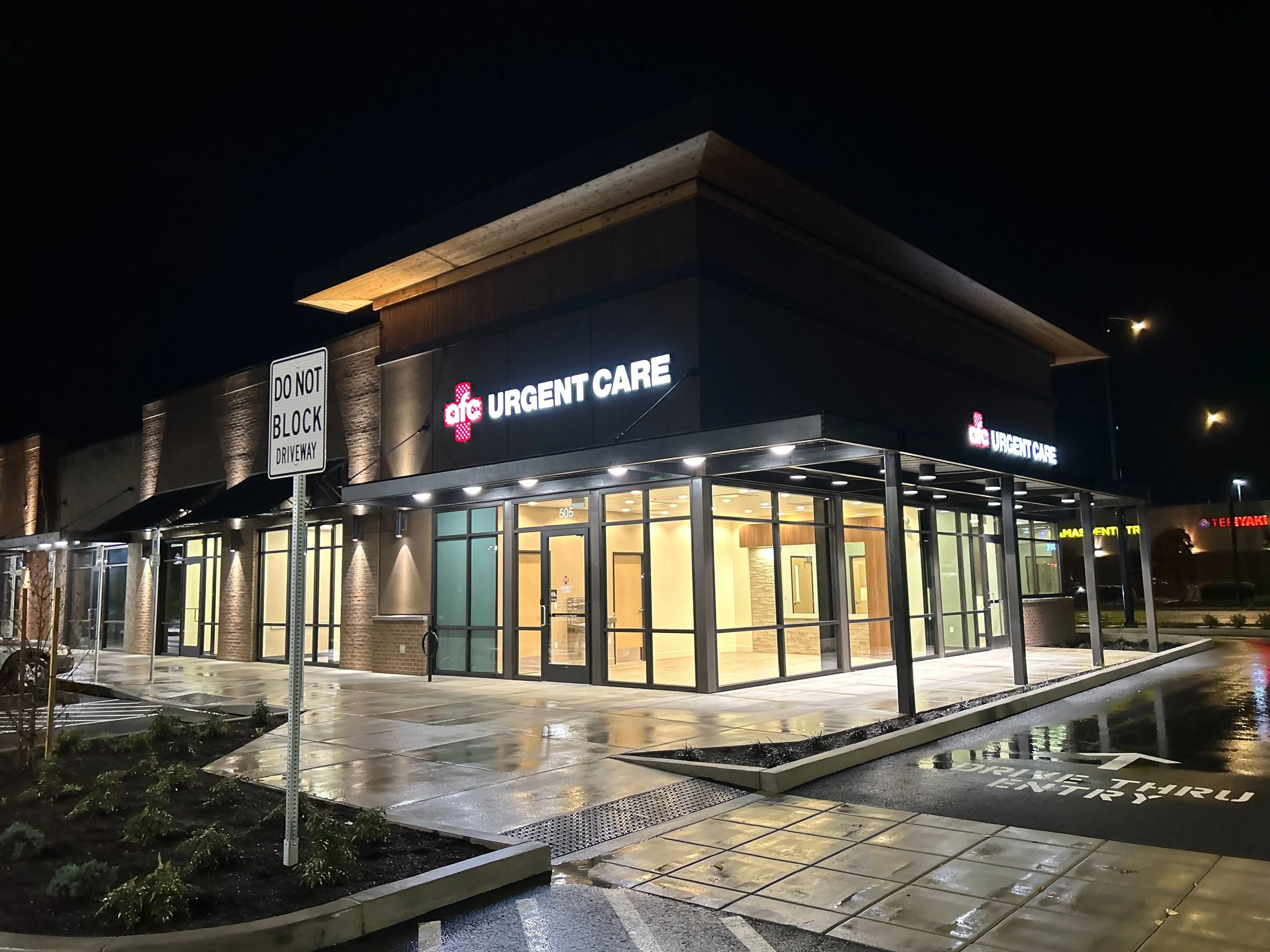 Nighttime exterior view of an urgent care center with illuminated signs that say 'AFG URGENT CARE'. The building has large glass windows, a covered entrance, and wet pavement reflecting the lights.