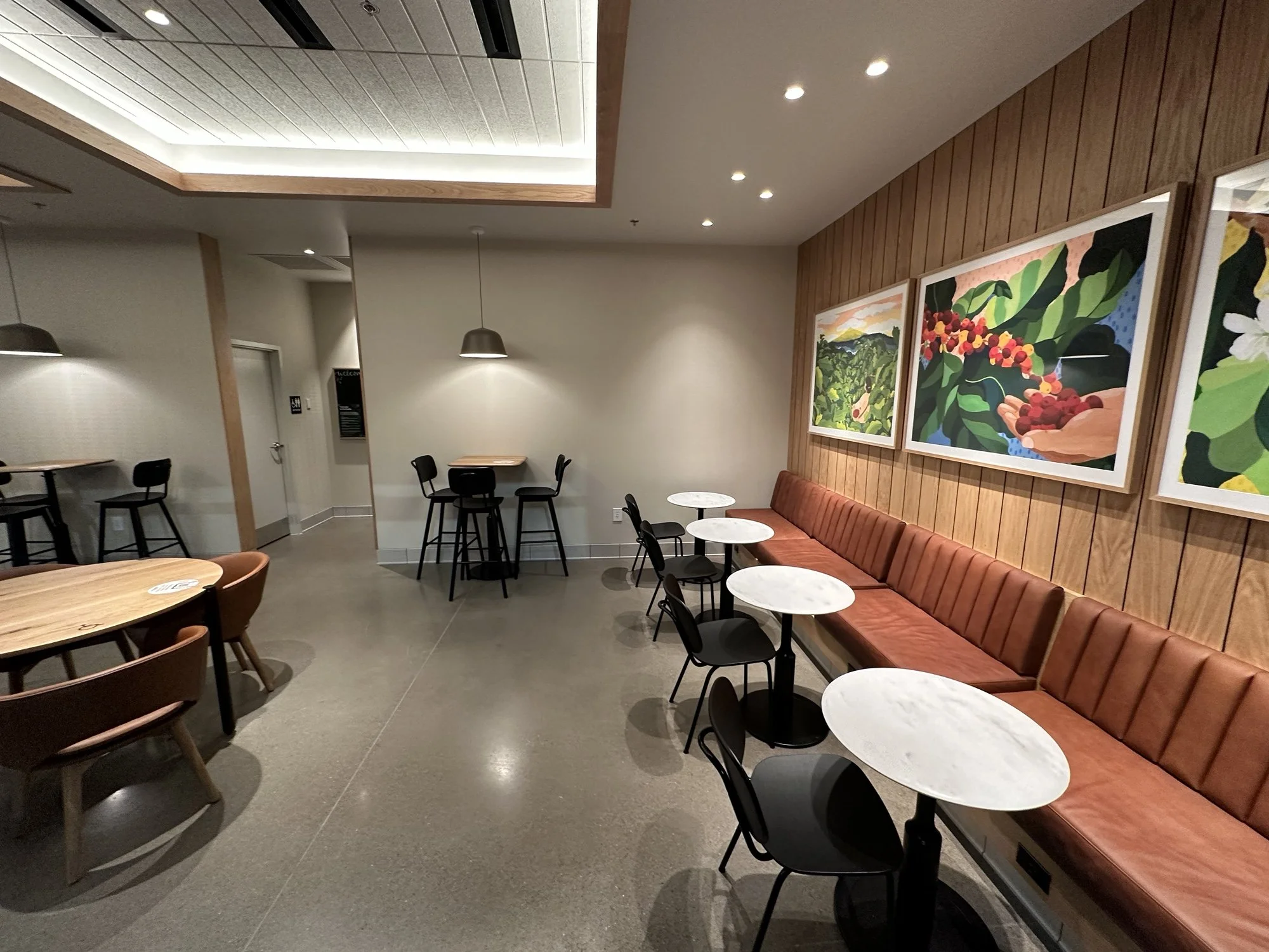 Empty cafe with round marble tables, black chairs, a long brown cushioned bench, and colorful artwork on a wood-paneled wall.