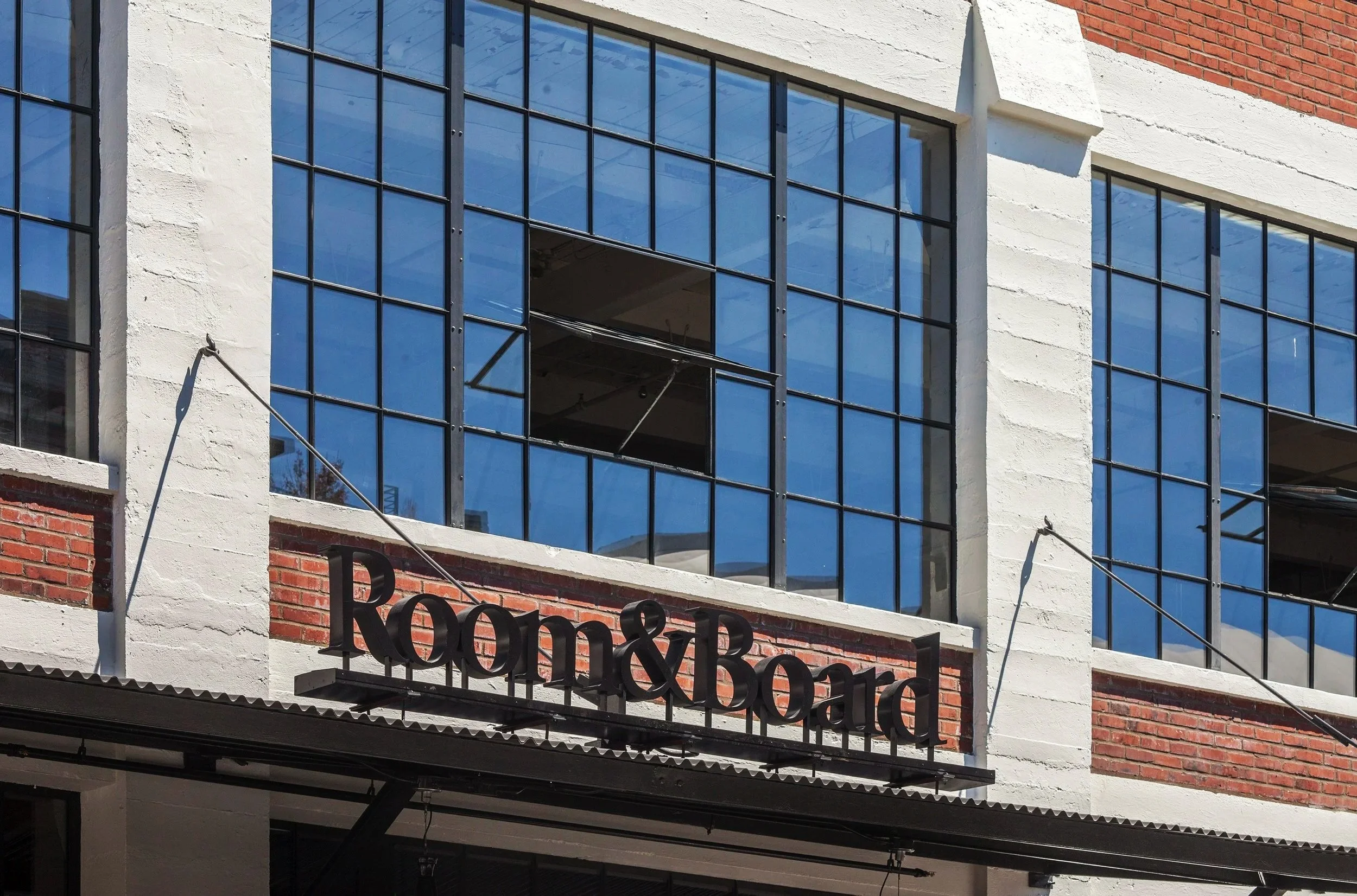 Exterior of a building with large blue-tinted windows, red brick accents, and a sign that reads 'Room & Board' in black letters.