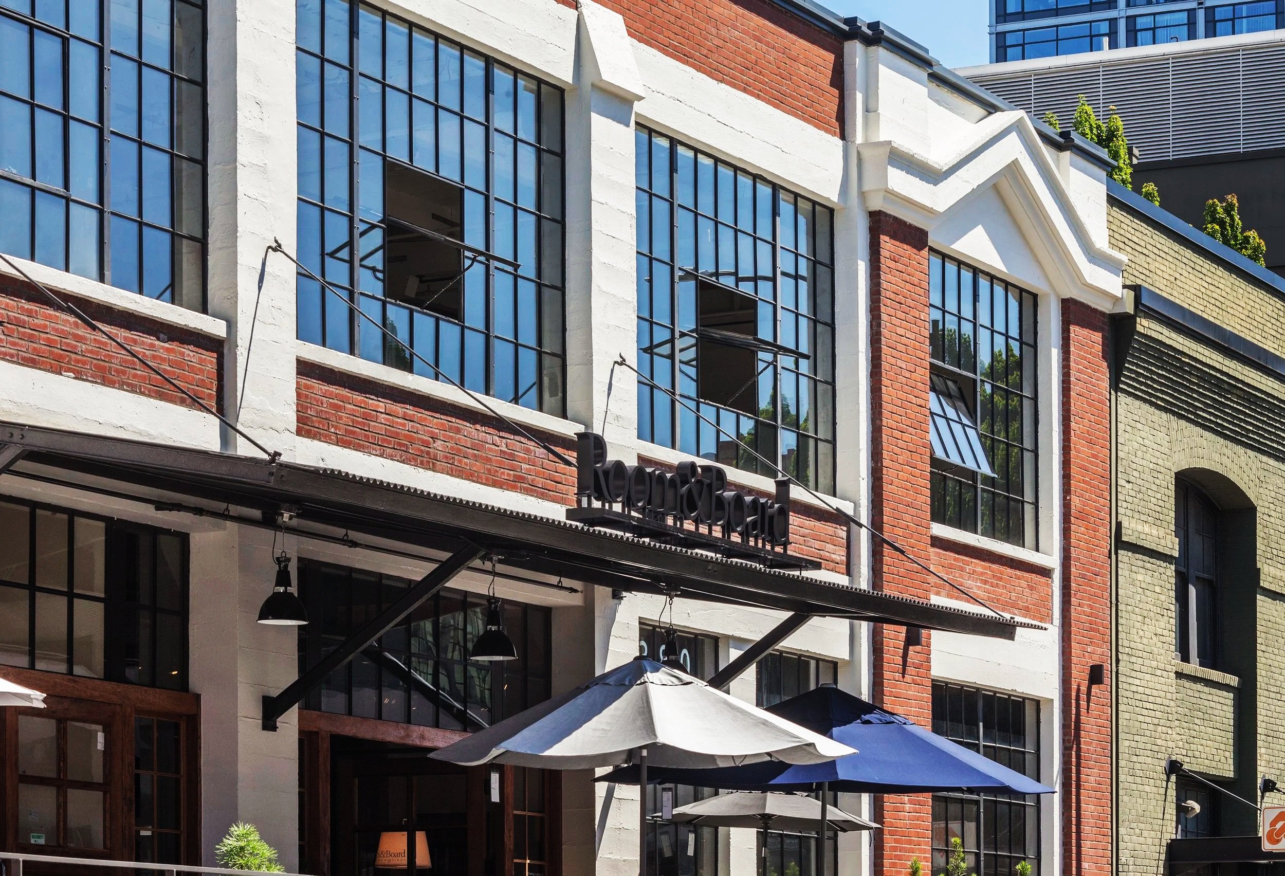 Exterior of a commercial building with large paned windows and a sign that reads 'Room & Board'. Outdoor seating with umbrellas is visible in the foreground.