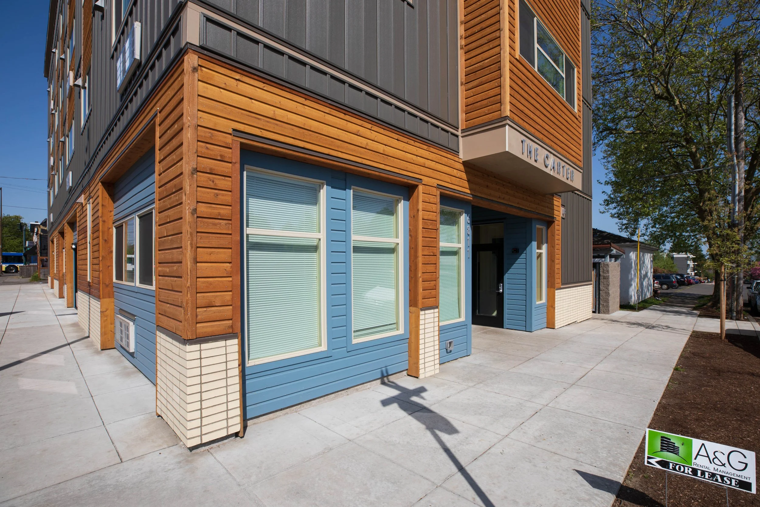 Modern multi-story building with wooden and blue siding exterior, multiple windows with blinds, sidewalk, and a for lease sign in the foreground reading "A&G For Lease." There is a tree with green foliage and parked cars in the background.