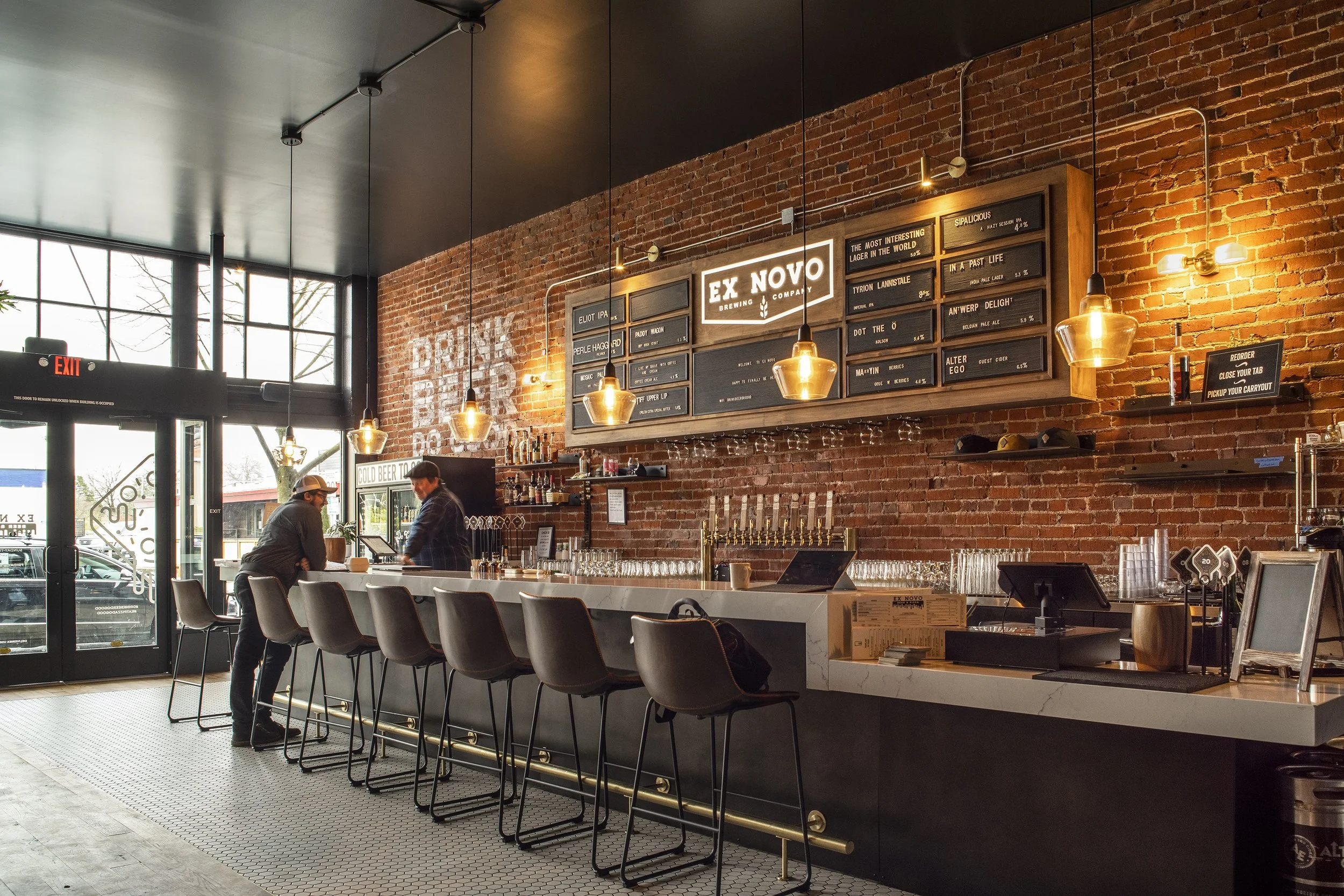 Inside a modern brewery or bar with a brick wall, large windows, and a white counter. Two people are seated at the bar, and another person is working behind it. There are hanging Edison-style light bulbs, a black chalkboard menu with various beers, a