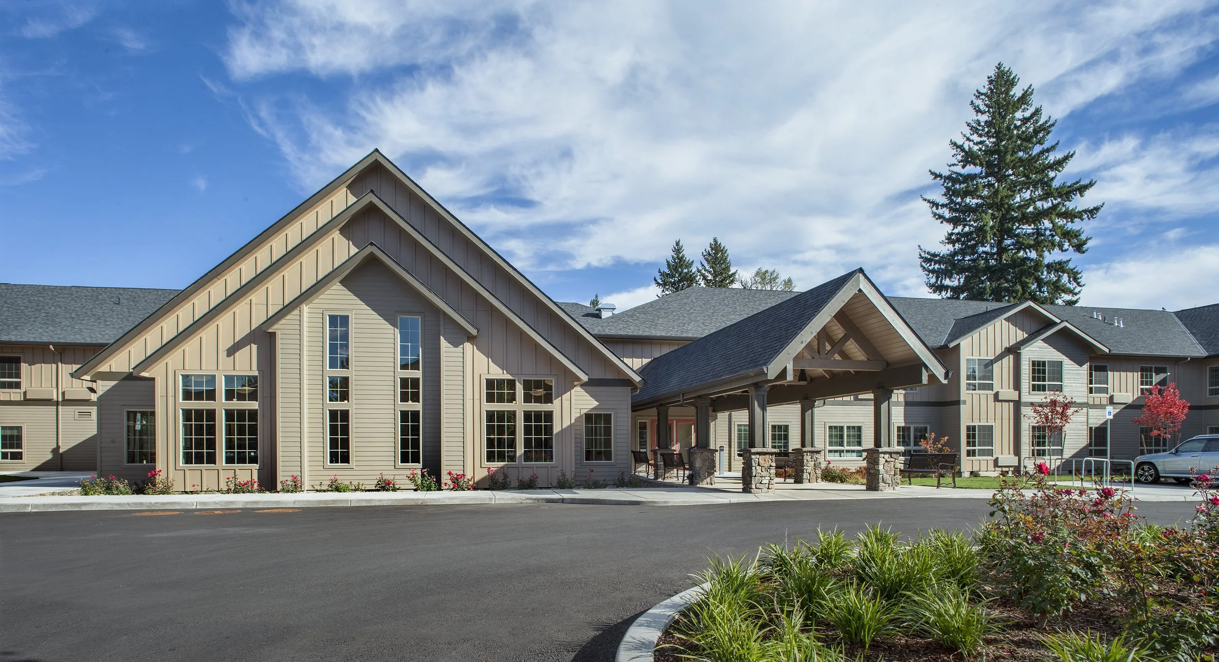 A modern residential building with beige siding and large windows, featuring a gabled roof and a covered entrance with stone columns, surrounded by a landscaped lawn, bushes, and trees under a partly cloudy sky.