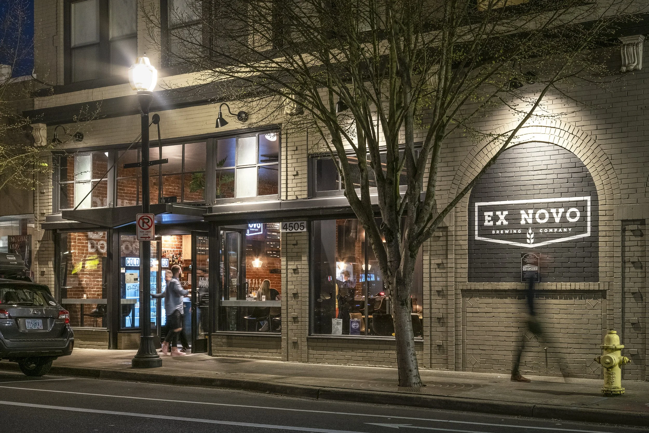 Nighttime view of a brewery named 'Ex Novo Brewing Co.' with illuminated sign on a brick building, a tree in front, and people inside visible through large windows.