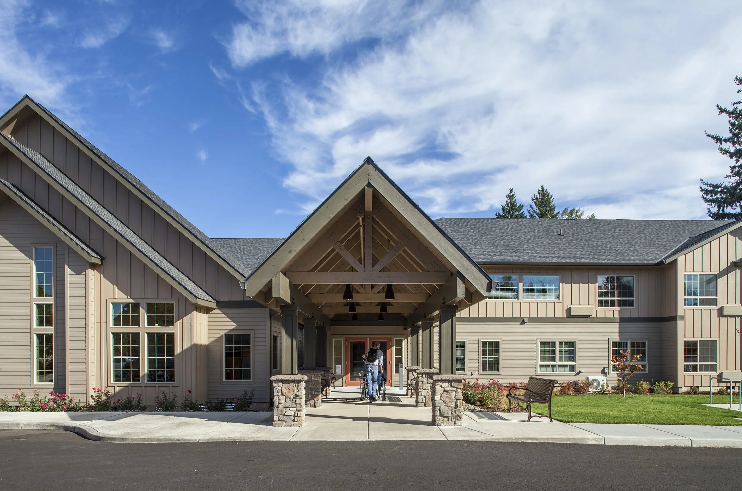 A modern, two-story residential building with a covered entrance, stone pillars, and landscaped front yard under a blue sky with scattered clouds.