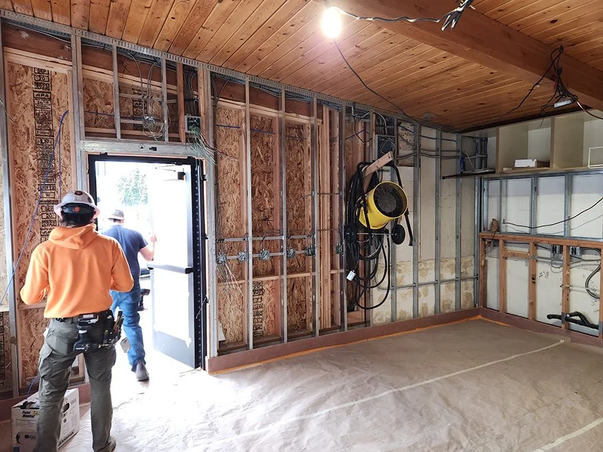 Construction workers working inside a room with exposed wall studs, wiring, and a wooden ceiling, as part of a building renovation or construction project.