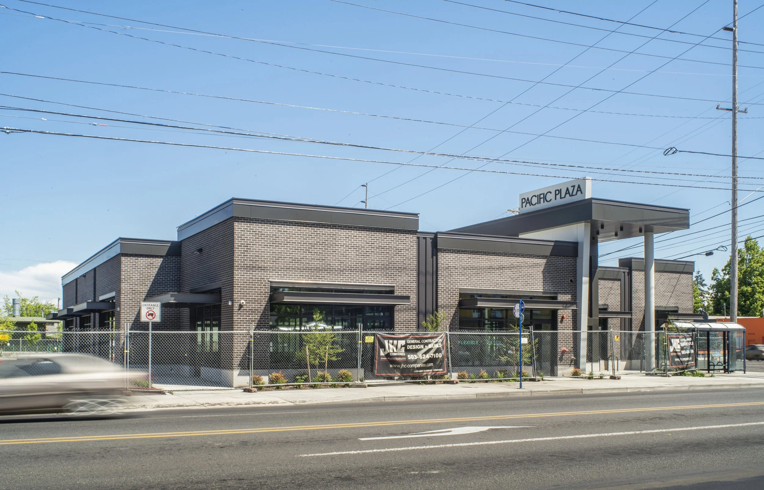 Modern brick building labeled Pacific Plaza, with a fenced construction area in front and a bus stop on the sidewalk, under a blue sky with power lines overhead.