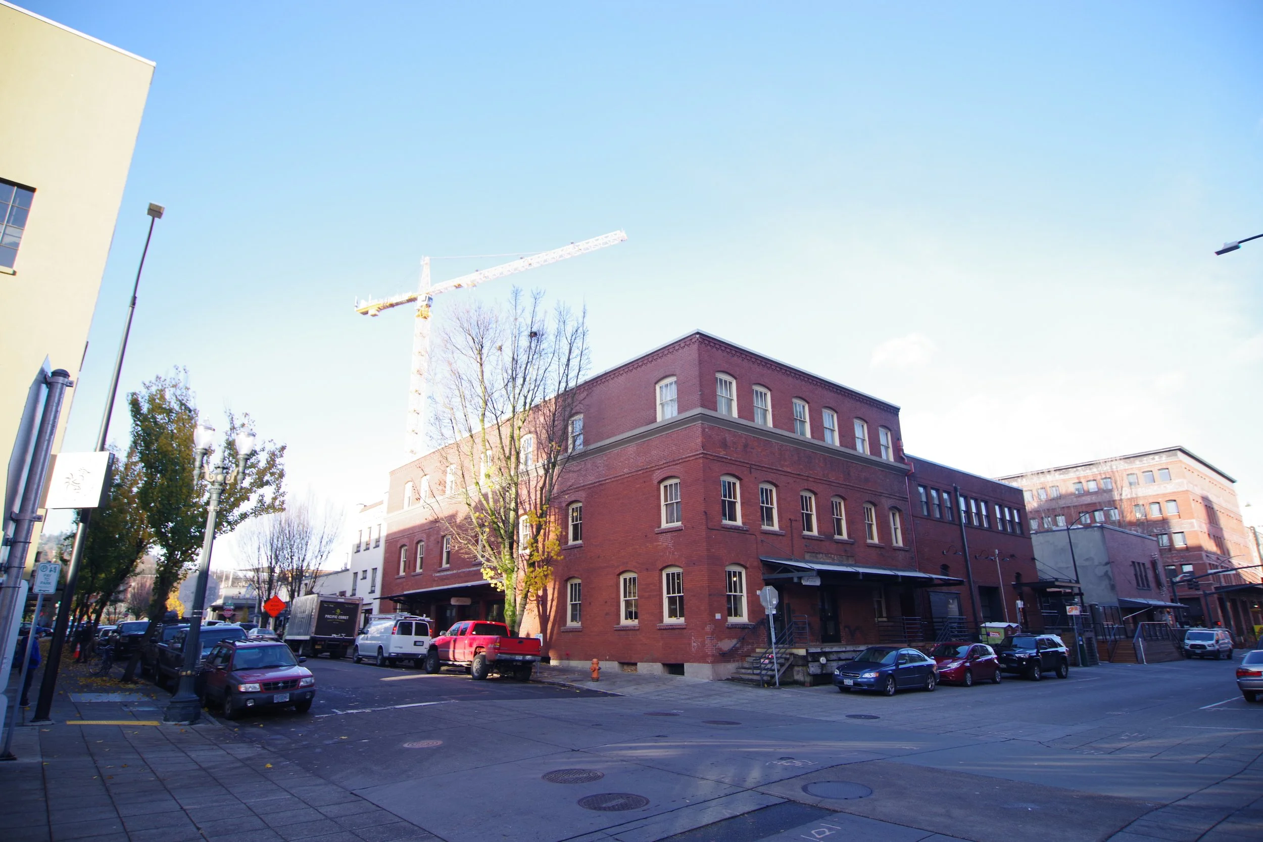 A street scene with parked cars, a brick building, leafless trees, and a construction crane in the background under a clear blue sky.