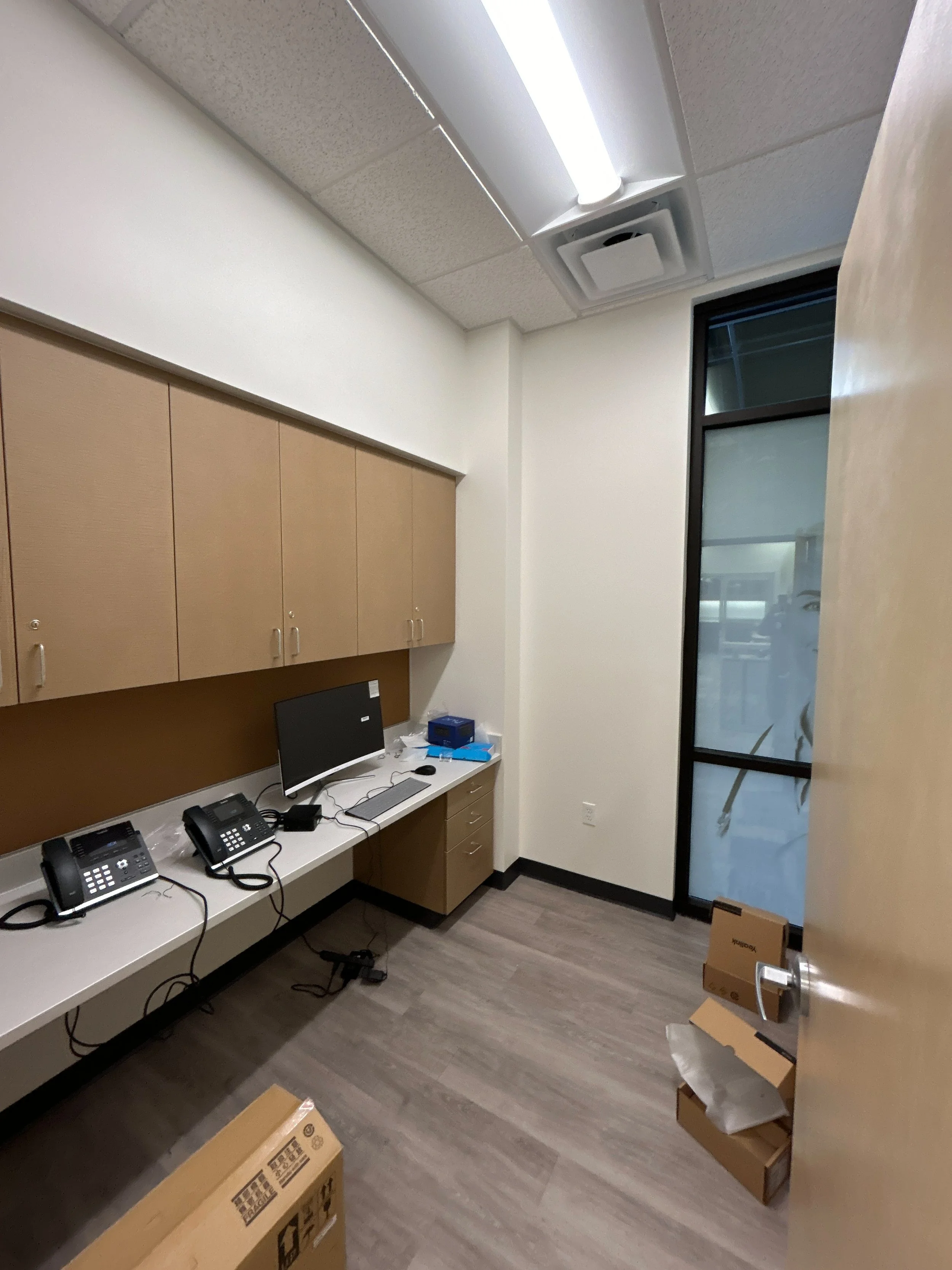 Empty office room with a desk, two phones, a monitor, a box, and some cardboard boxes on the floor near the door.