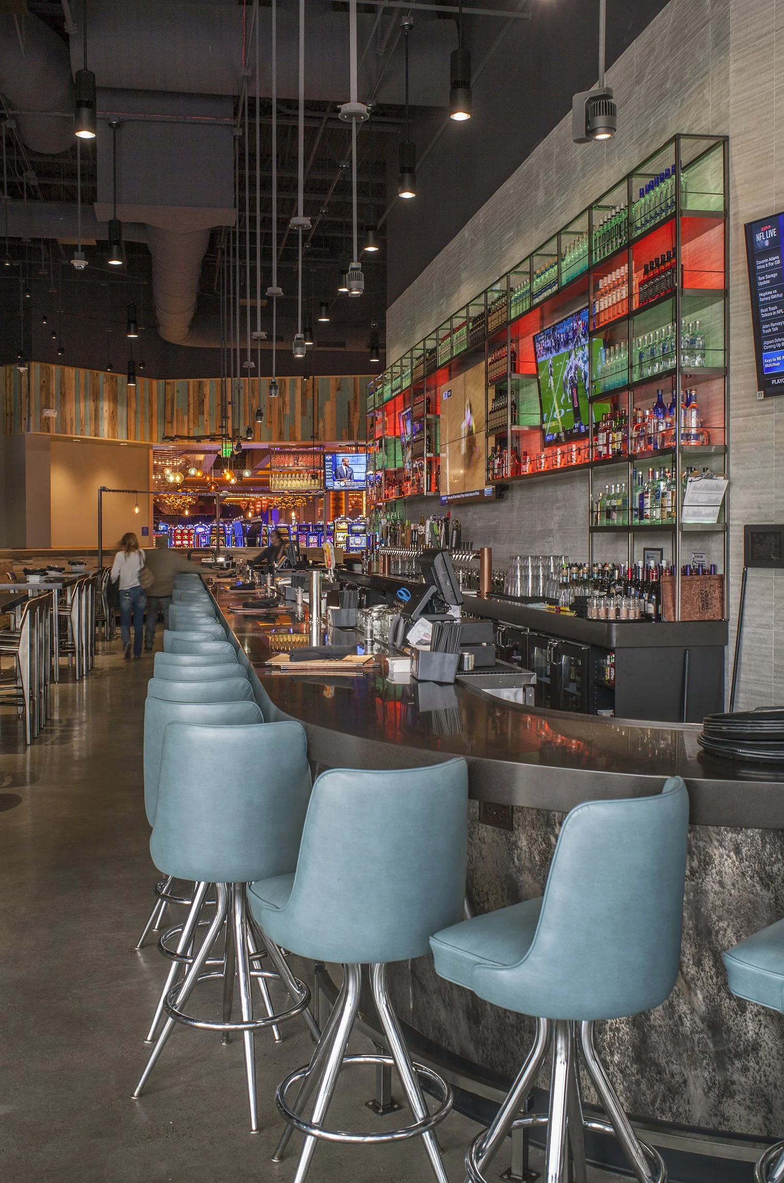 Interior of a modern bar with high-top teal chairs, a black curved bar counter, colorful shelving with liquor bottles, and multiple TVs showing sports.