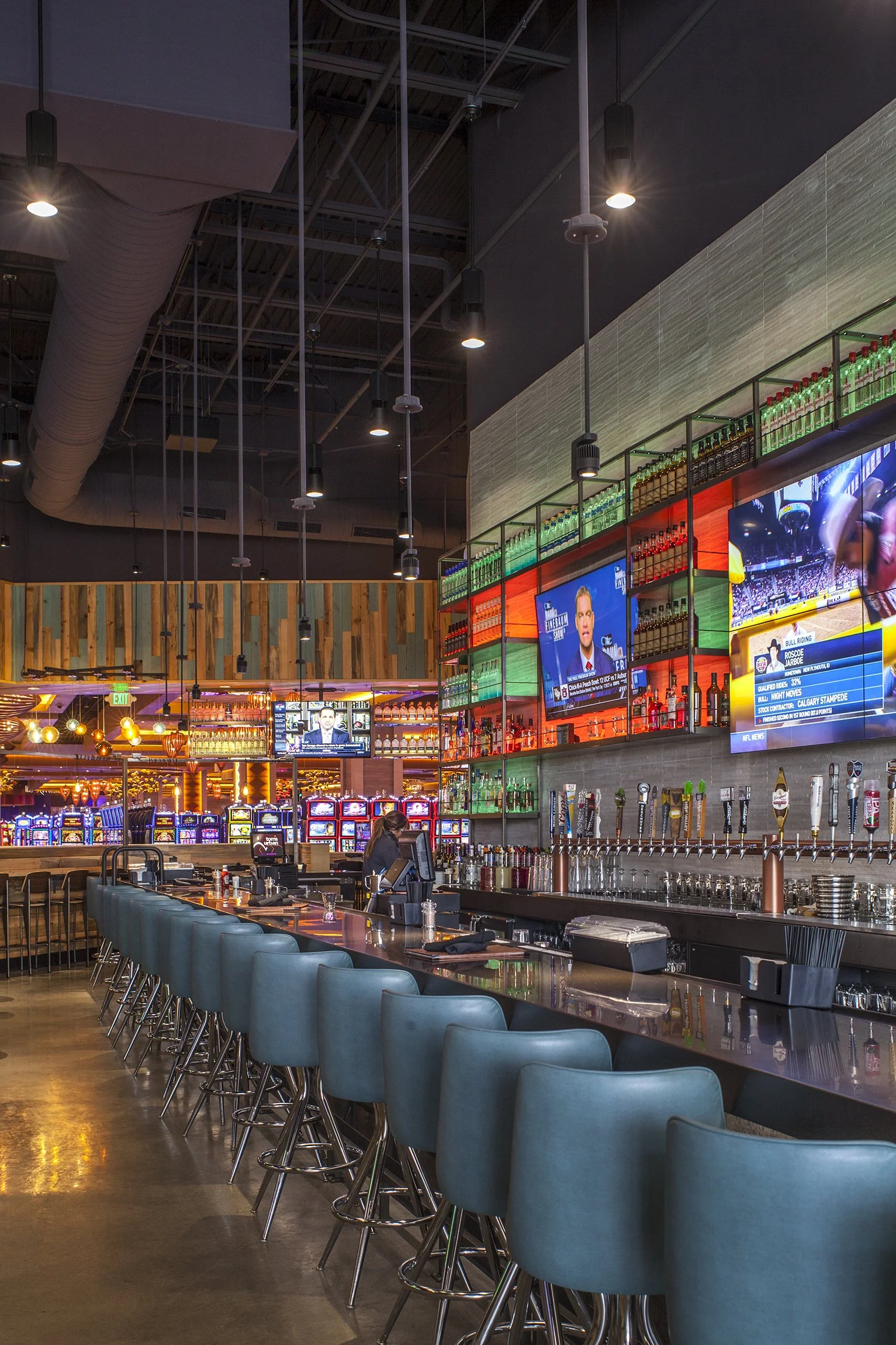 Interior of a modern bar with teal bar stools, multiple television screens, colorful lighting, and a row of beer taps.