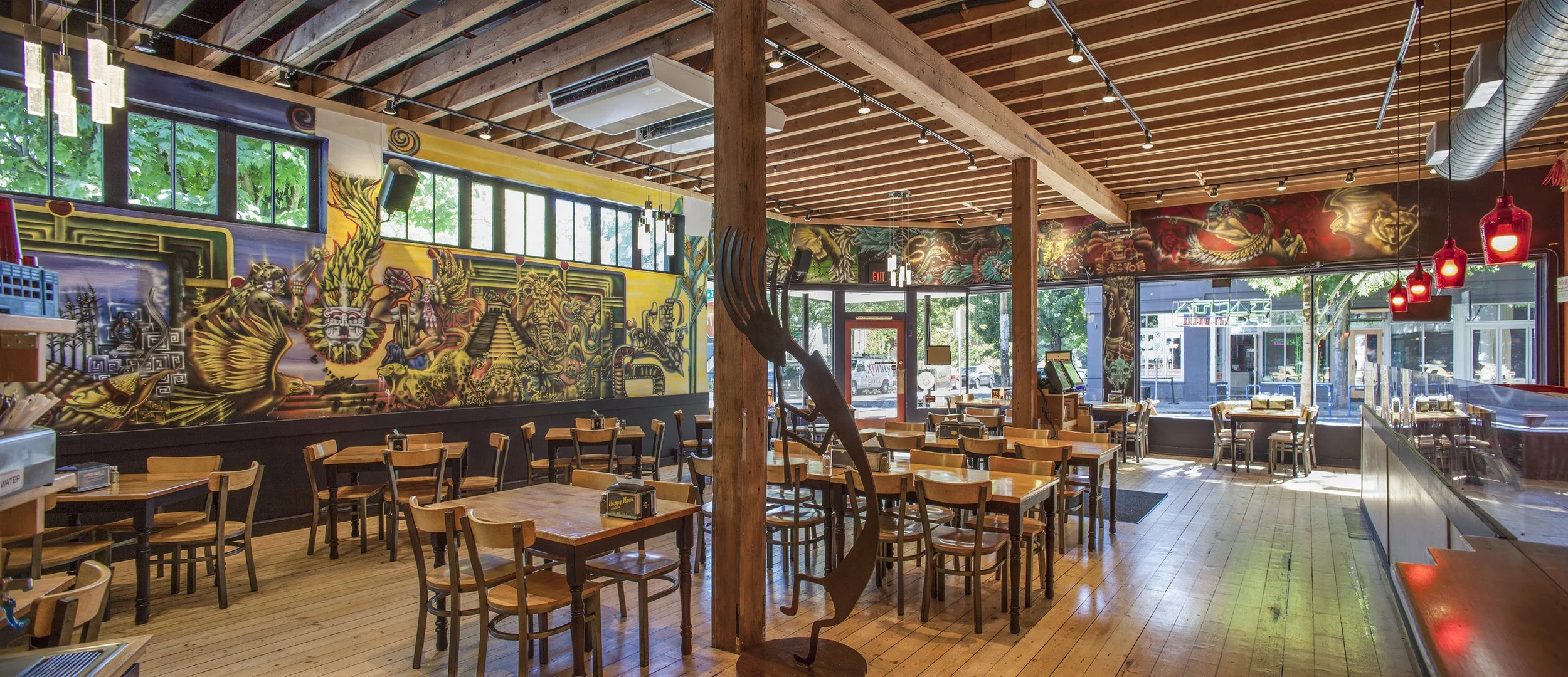 Interior of a restaurant with wooden floors, tables, and chairs. Colorful mural with Aztec motifs on the wall, large glass windows letting in natural light, and red pendant lights hanging from the ceiling. Wooden beams and modern decor.