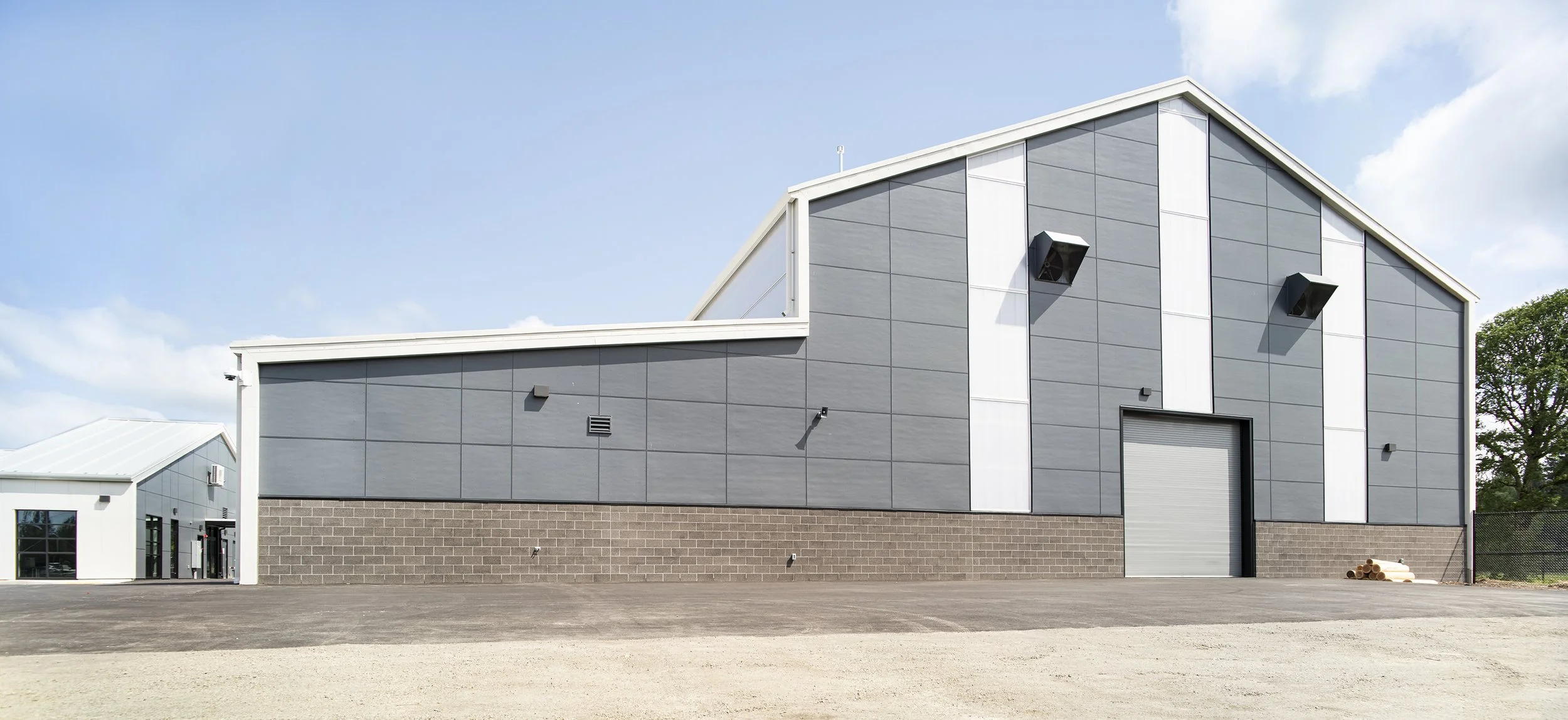 A modern industrial warehouse building with gray and white exterior panels, large garage door, and black ventilation speakers mounted on the walls. The area in front is paved, with some pipes stacked near the right corner, under a partly cloudy sky.