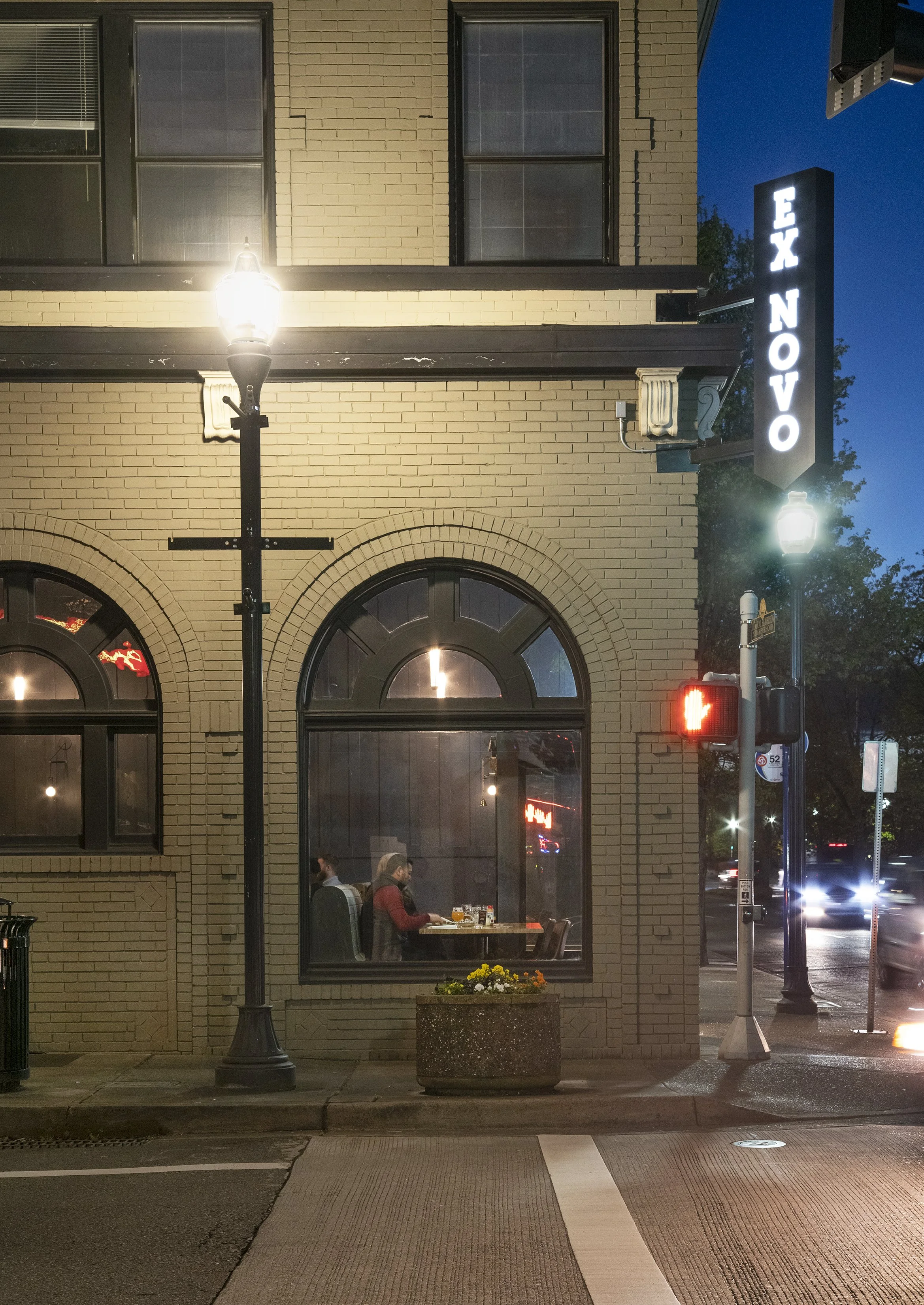 Nighttime street scene outside a brick building with a lit sign reading 'EX NOVO,' a pedestrian crossing light, and a window showing people dining inside.