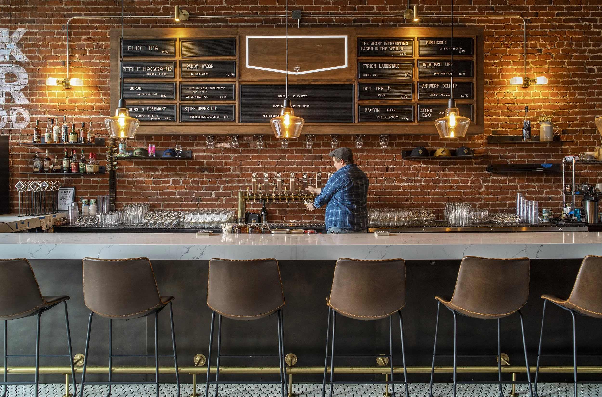 Interior of a modern bar with a white marble counter, set against a red brick wall. A bartender in a blue plaid shirt stands behind the counter, handling glasses and taps. Above, blackboards display drink names and percentages, framed with wood, unde