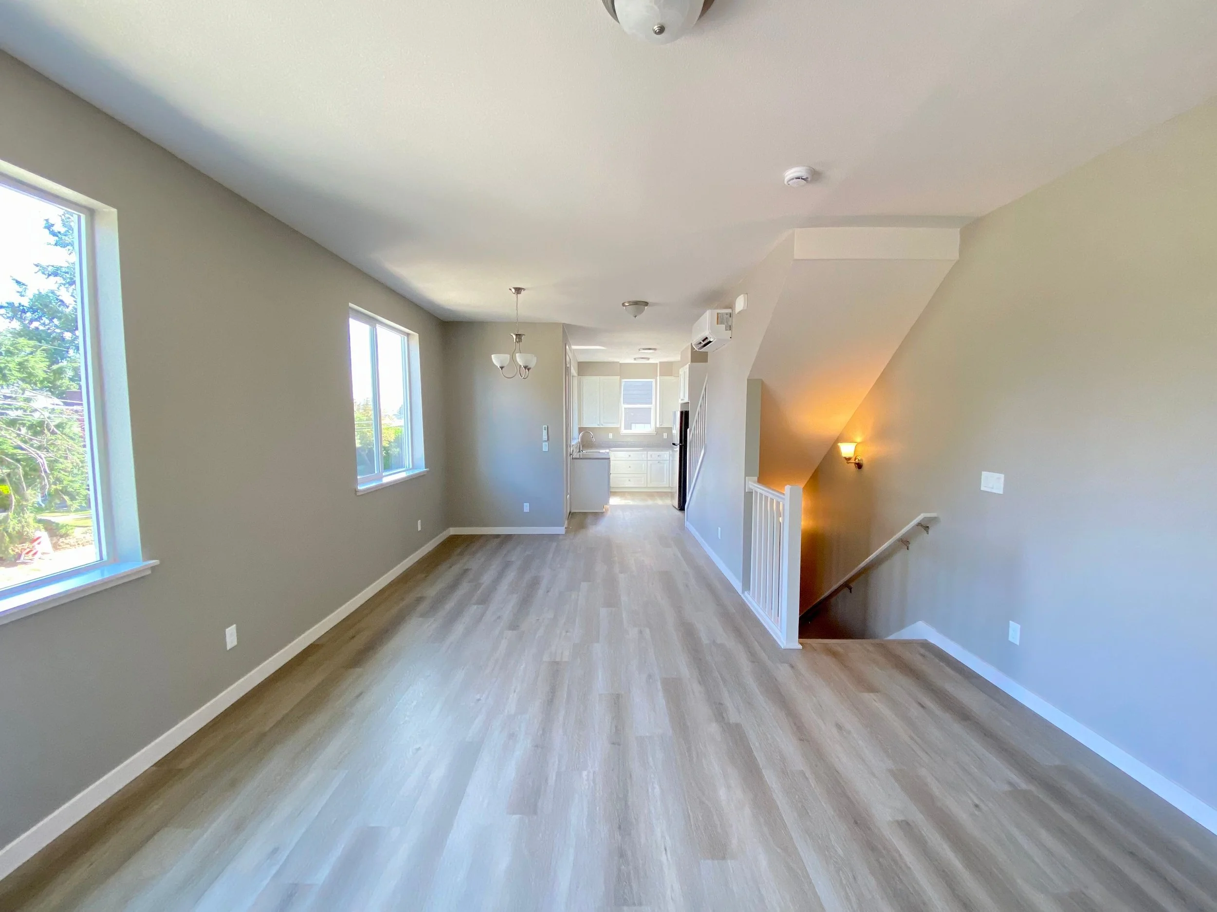 Empty living room with large windows, light wood flooring, beige walls, and a staircase leading downstairs. Kitchen visible in the background.