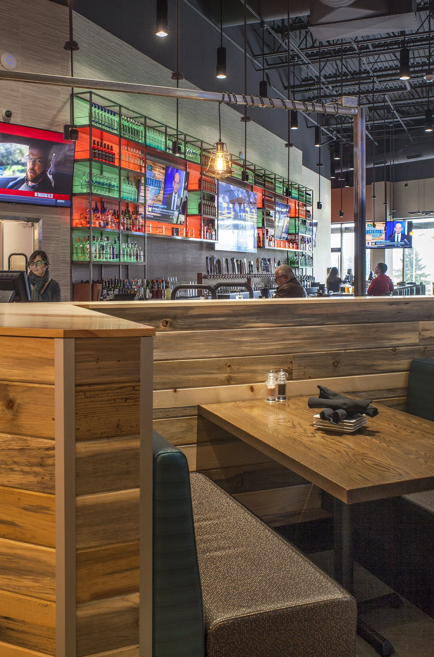 Interior of a modern bar or restaurant with a wooden partition, a table set with napkins and salt and pepper shakers, a bar in the background with multiple taps, and TV screens displaying news and show.