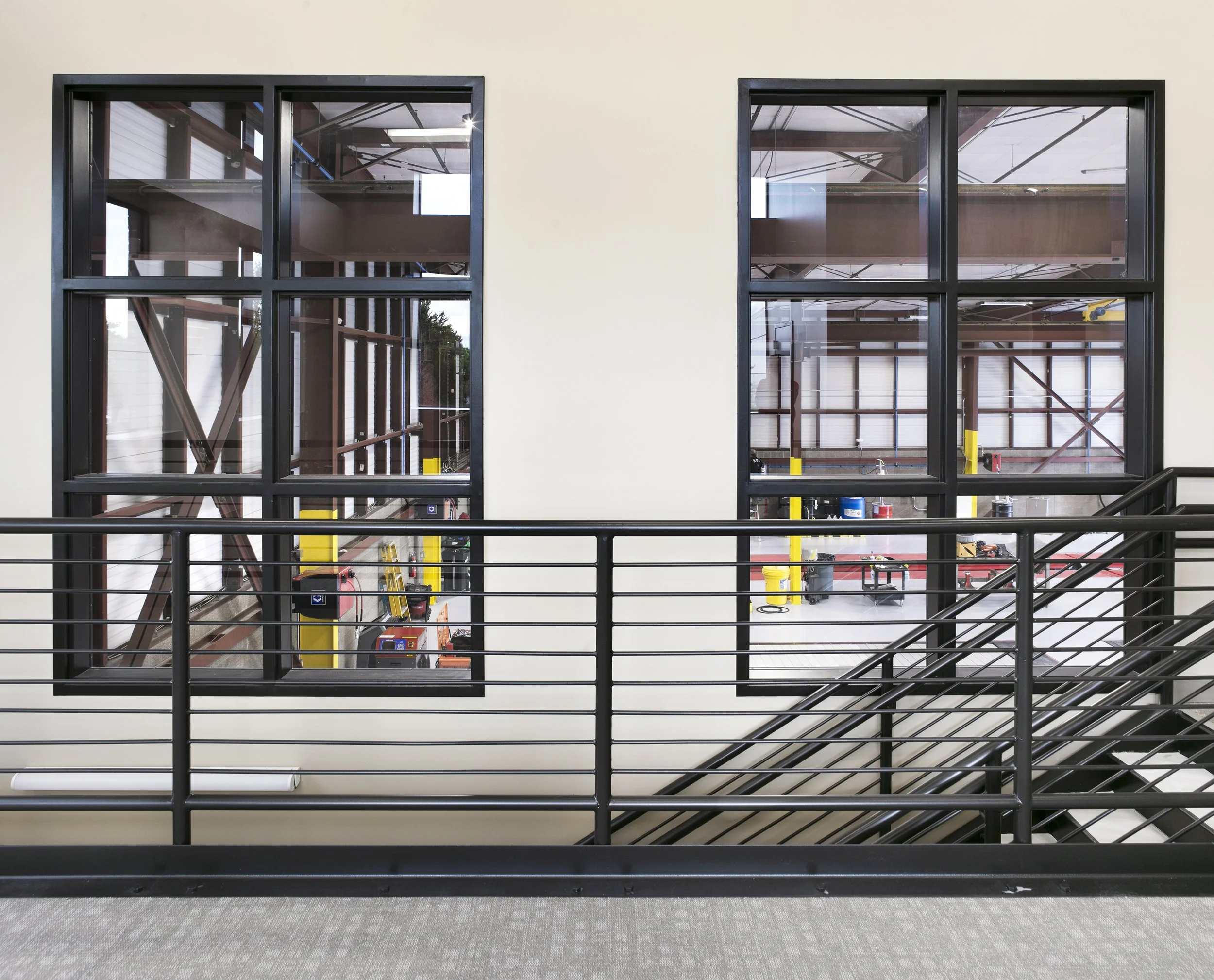 Interior of a building under construction, viewed from a hallway with a metal railing and staircase, showing large windows revealing a partially built structure and construction tools inside.