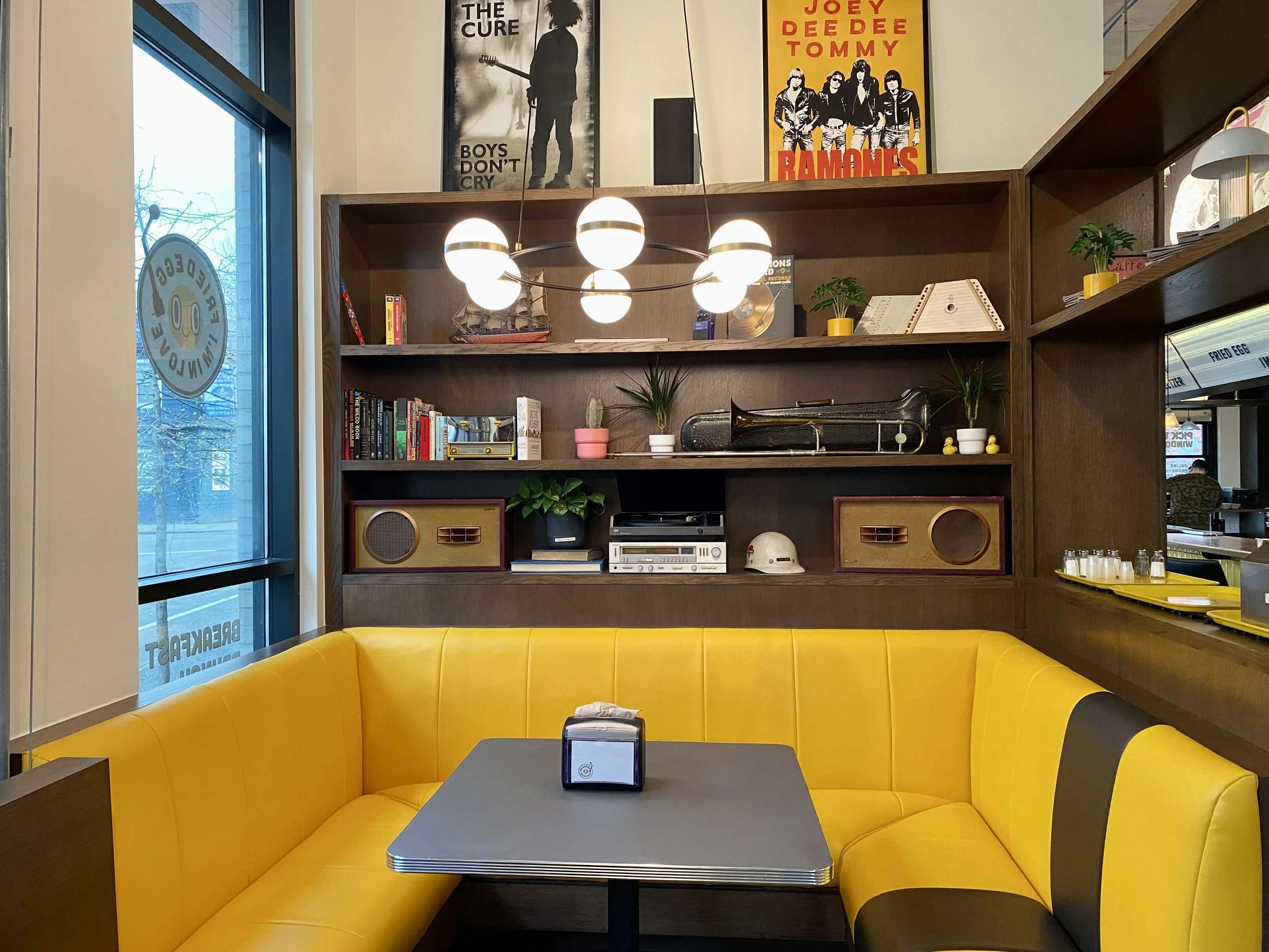 A cozy corner in a restaurant or cafe featuring a bright yellow cushioned booth, a dark gray table with a tissue holder, and a wooden shelf with books, decorative items, and posters on the wall. There is a window with a sticker and a view of the outs