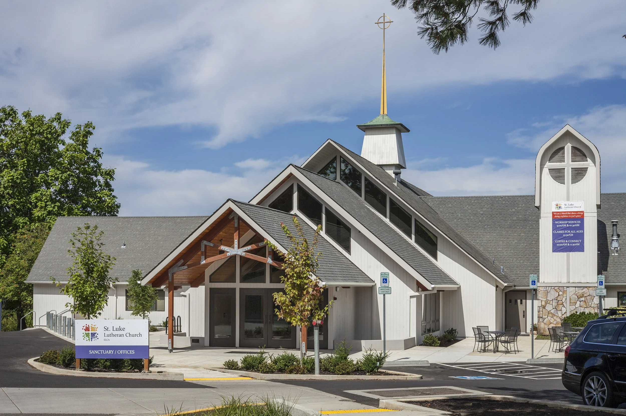Front view of St. Luke Lutheran Church and its sanctuary and offices, with a parking lot in front, trees, and a partly cloudy sky.