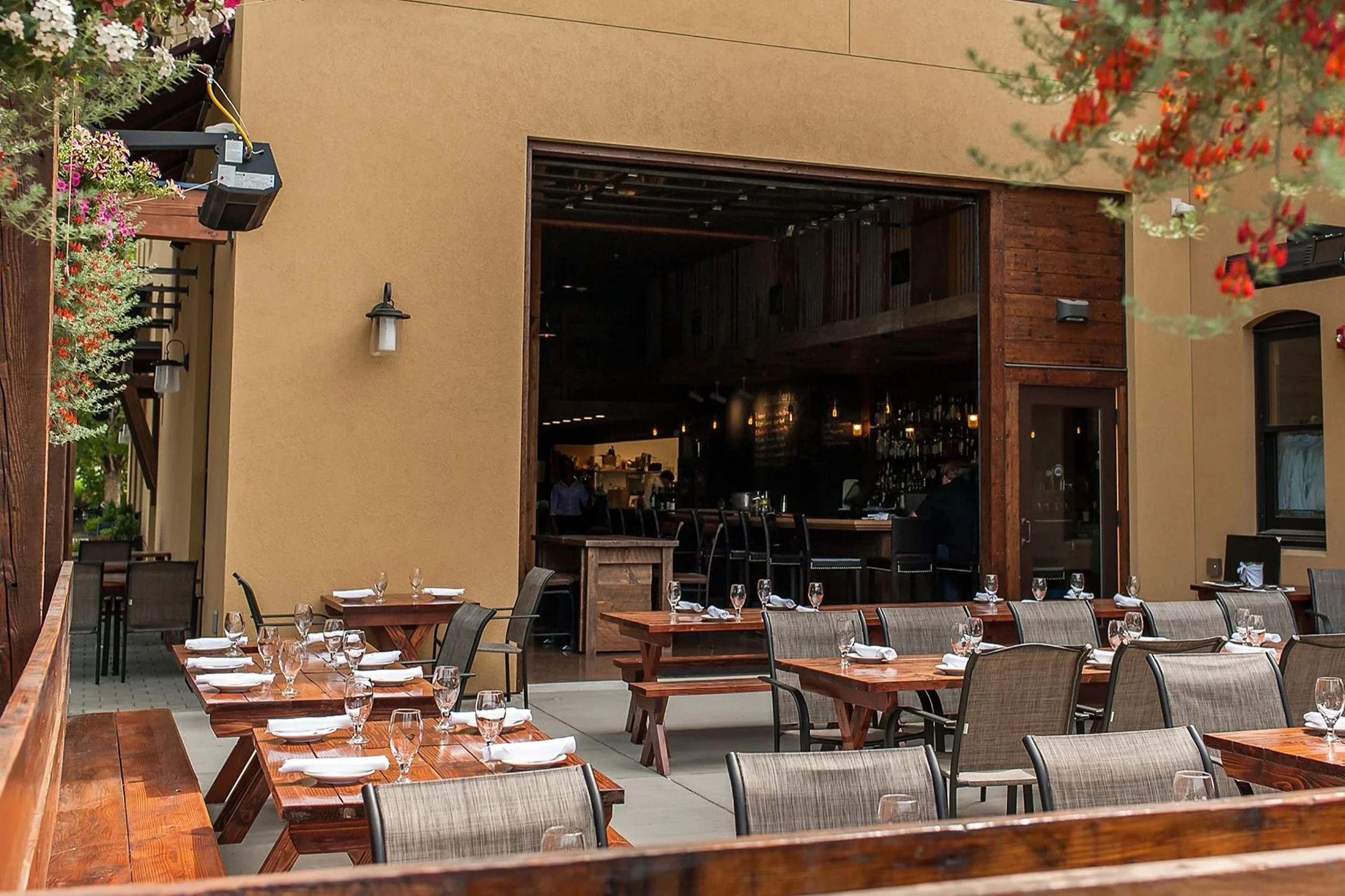 Outdoor patio of a restaurant with wooden tables set with glasses and napkins, with a view into an indoor bar area through an open doorway.