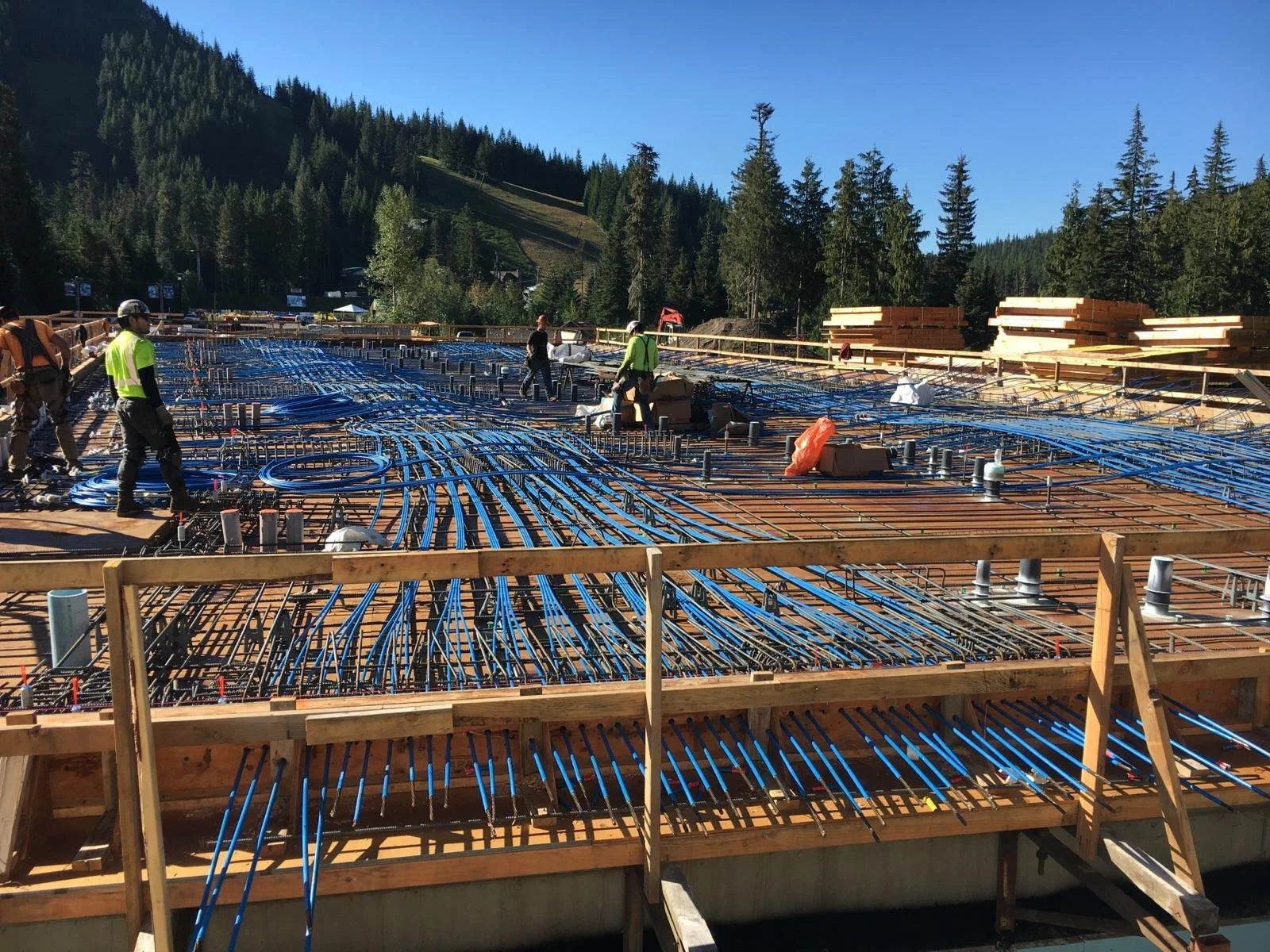 Construction workers install electrical wires and plumbing on a building's foundation with wooden framing, surrounded by treetops and mountains under a clear blue sky.