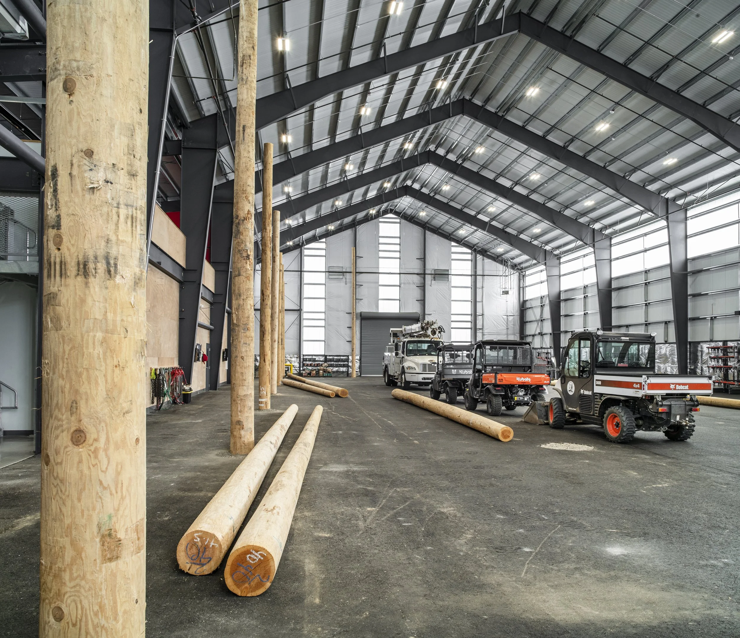 Interior of a large industrial warehouse with wooden logs, utility vehicles, and construction equipment.