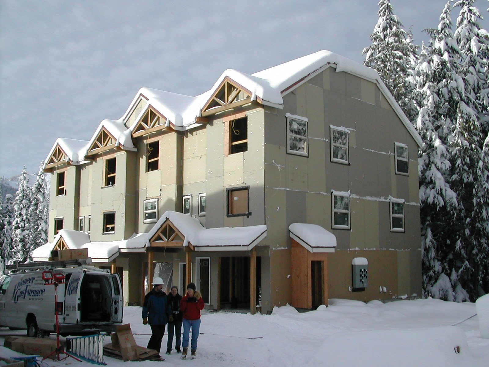 A three-story house under construction with snow on its roof and surrounding ground, and trees covered in snow in the background. Three people wearing winter clothing and hard hats are standing in front of the house, along with a van parked nearby.