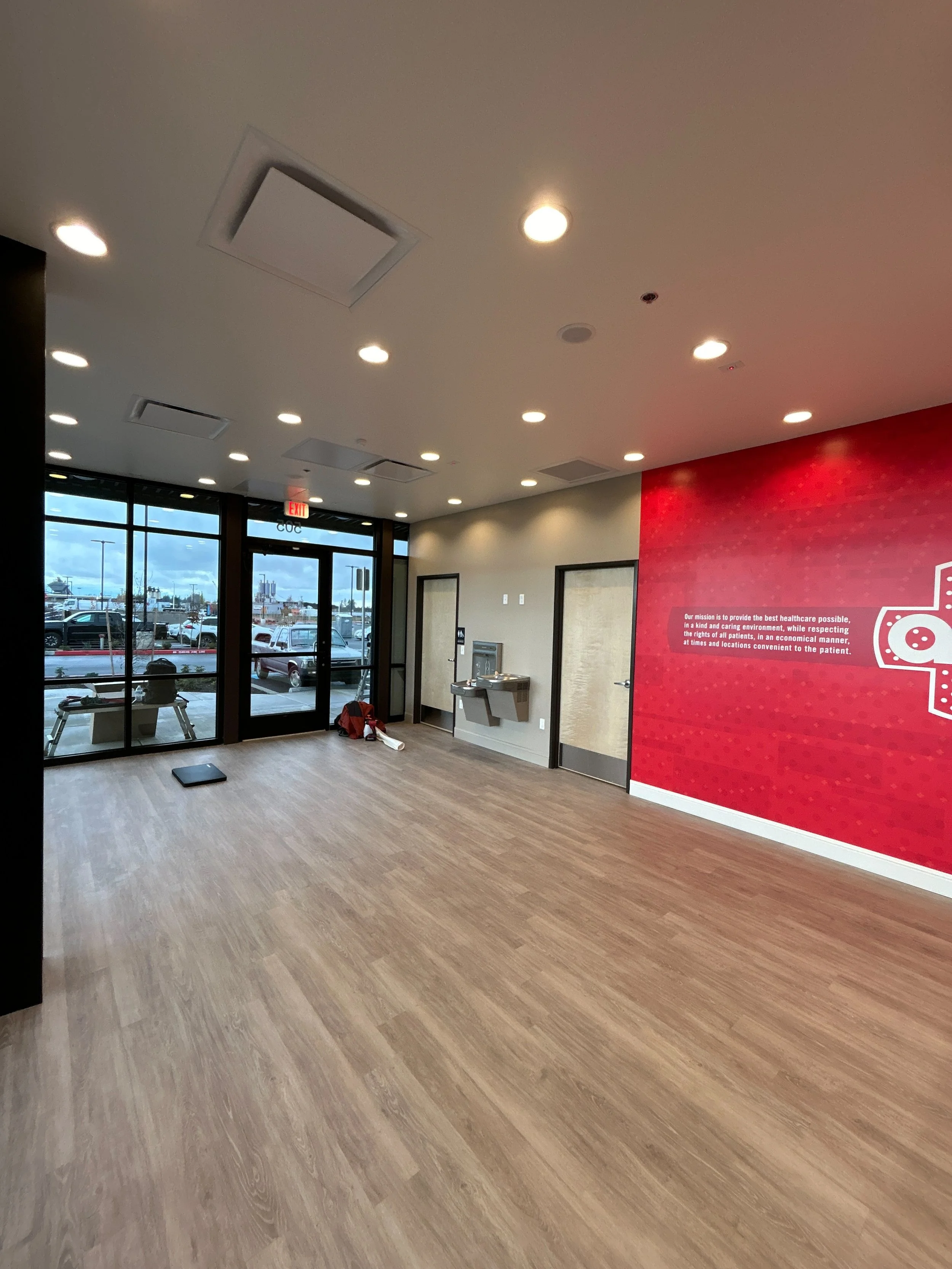 Empty room in a healthcare facility with wooden floor, glass doors, red wall with healthcare message, water fountain, and outdoor parking lot visible through glass