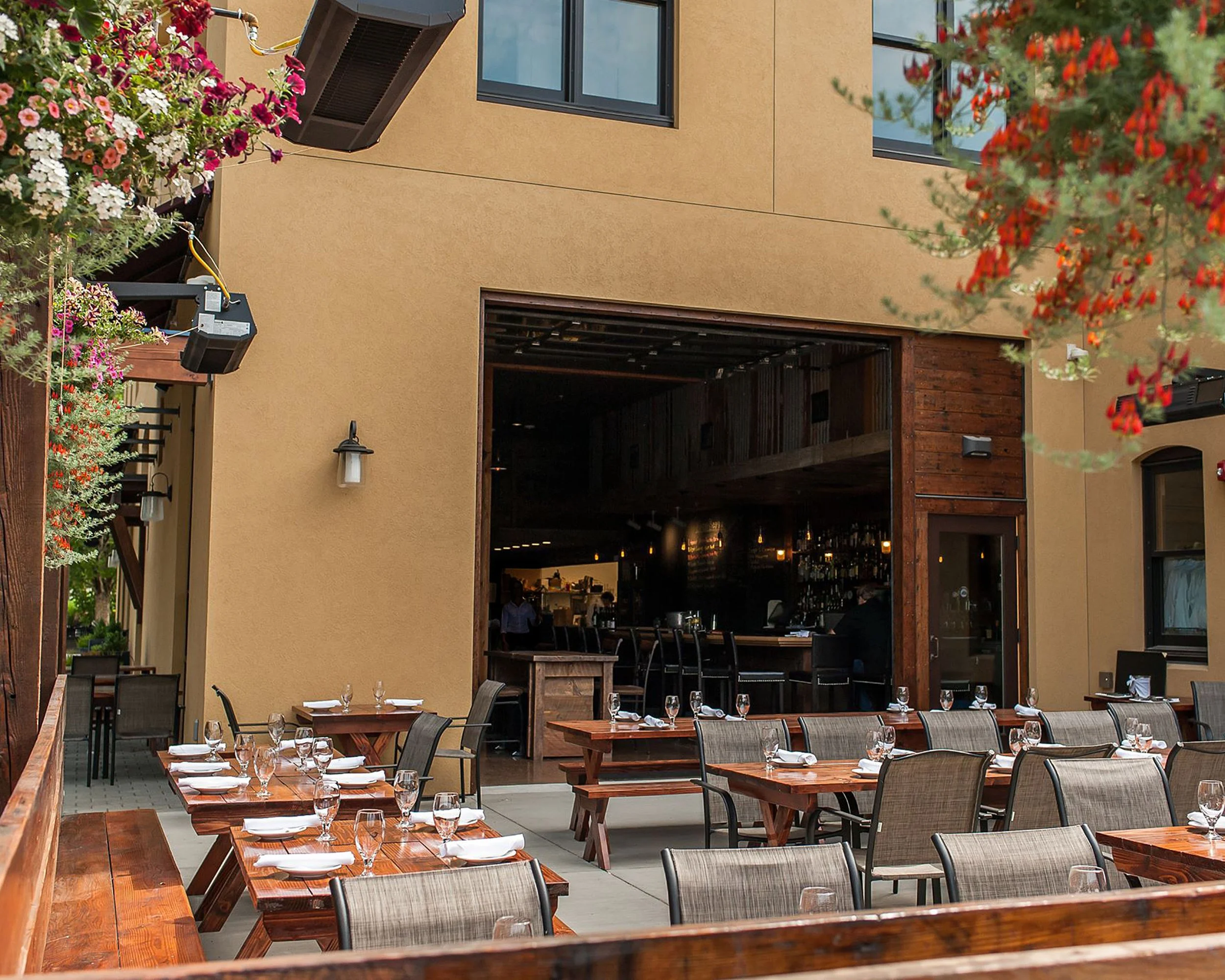 An outdoor dining area with wooden tables set with white plates, silverware, and glasses, surrounded by chairs with beige cushions. The area is next to a building with a yellowish exterior wall, some hanging flower baskets, and a large open window re