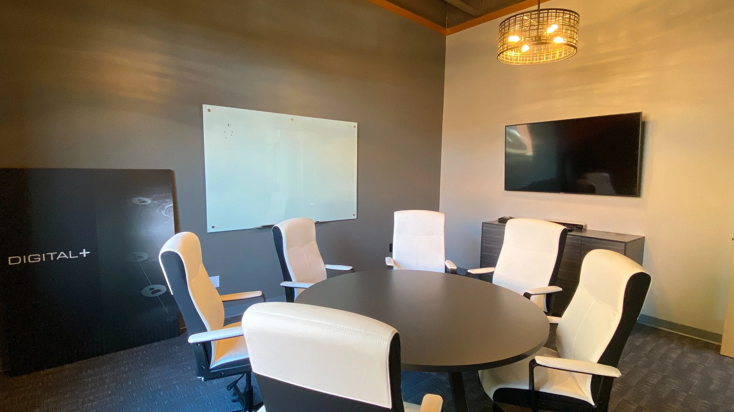 A conference room with a round black table surrounded by eight beige and black office chairs. A flat-screen TV is mounted on the wall on the right side, underneath a modern pendant light fixture. There is a large whiteboard on the grey wall in the ba