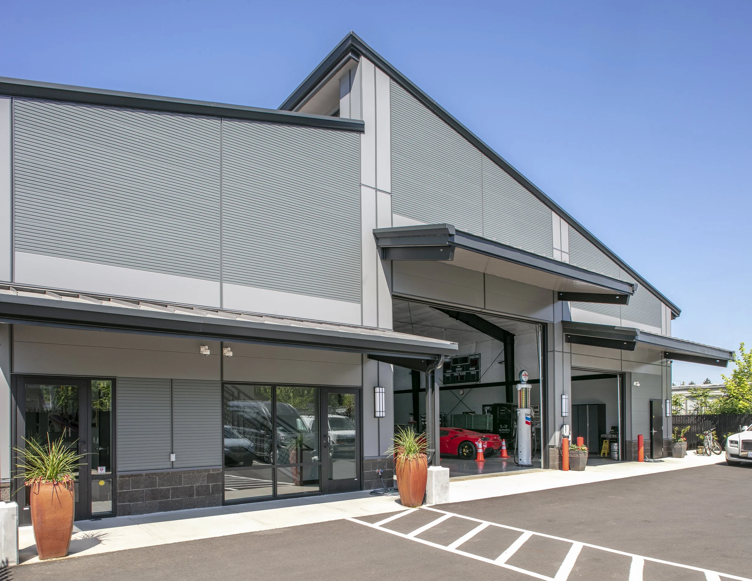 Modern commercial building with a parking lot, featuring plants in large pots, garage space with a red sports car, and blue sky.