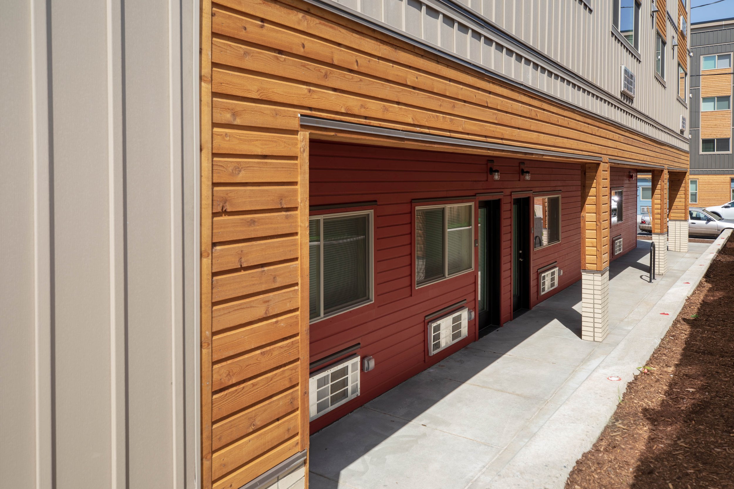Exterior view of a modern apartment building with wooden siding, windows, air conditioning units, and a sidewalk walkway.
