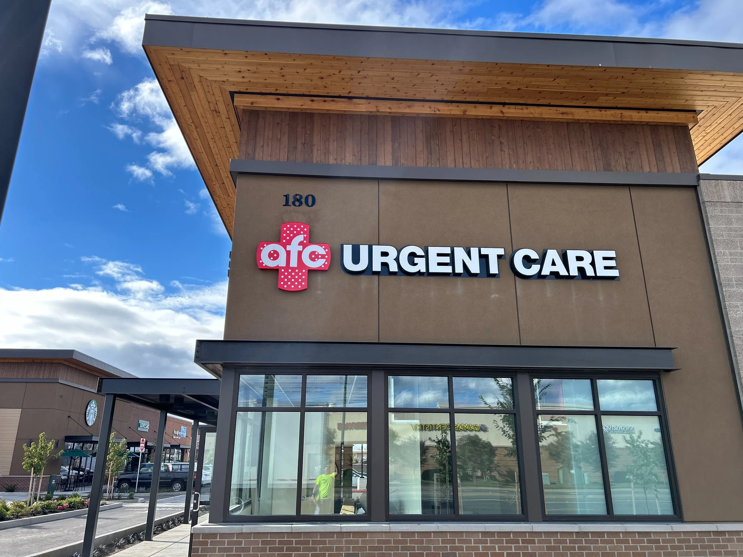 Exterior of an AFC Urgent Care clinic with a sign on a tan building, featuring the AFC logo with a red cross and white letters, and the words "URGENT CARE" in white above large glass windows, under a partly cloudy sky.