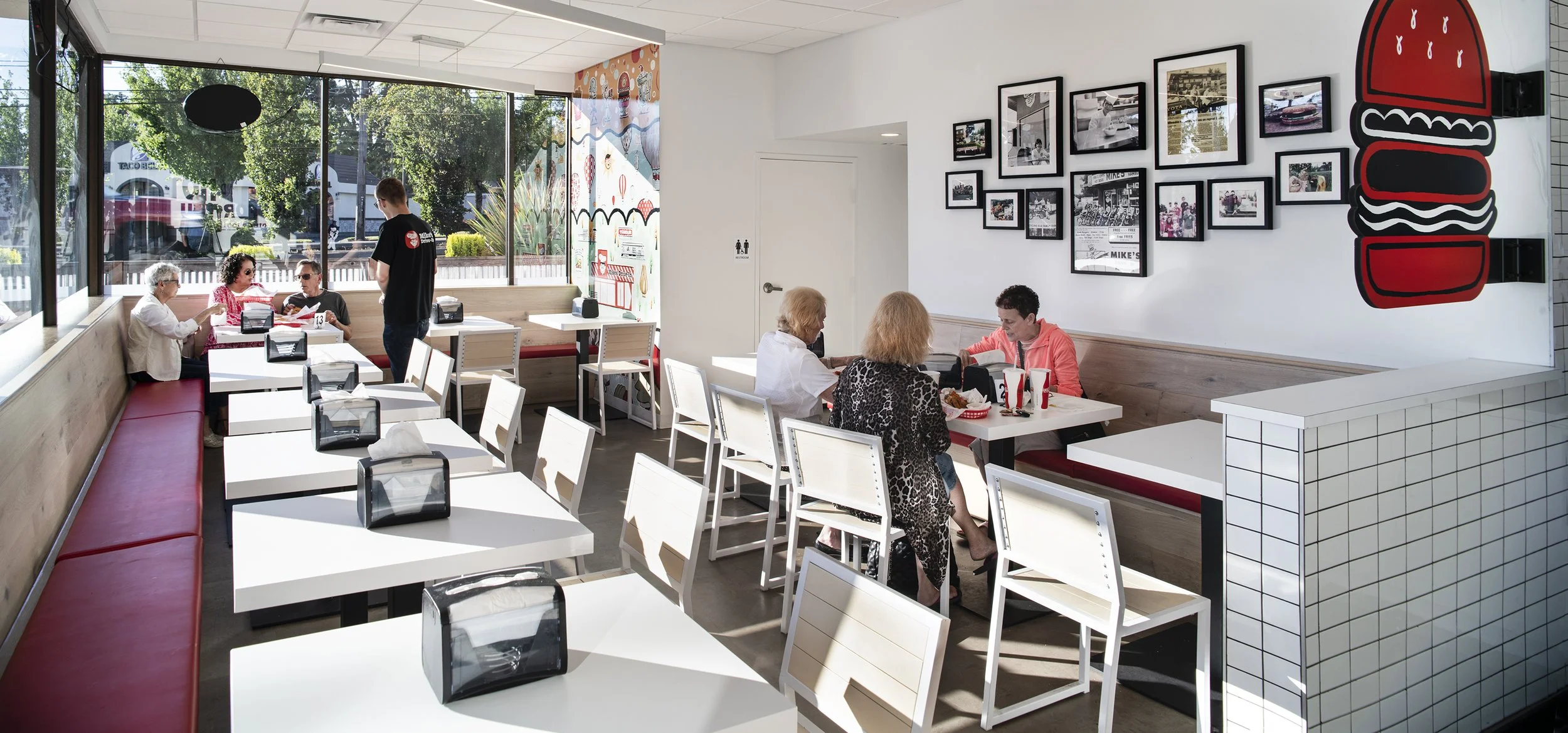 Interior of a casual restaurant with white tables, white chairs, and red cushioned bench seating. Five women are seated at tables, eating and talking, with a server attending one table. Large floor-to-ceiling windows let in natural light, and framed 