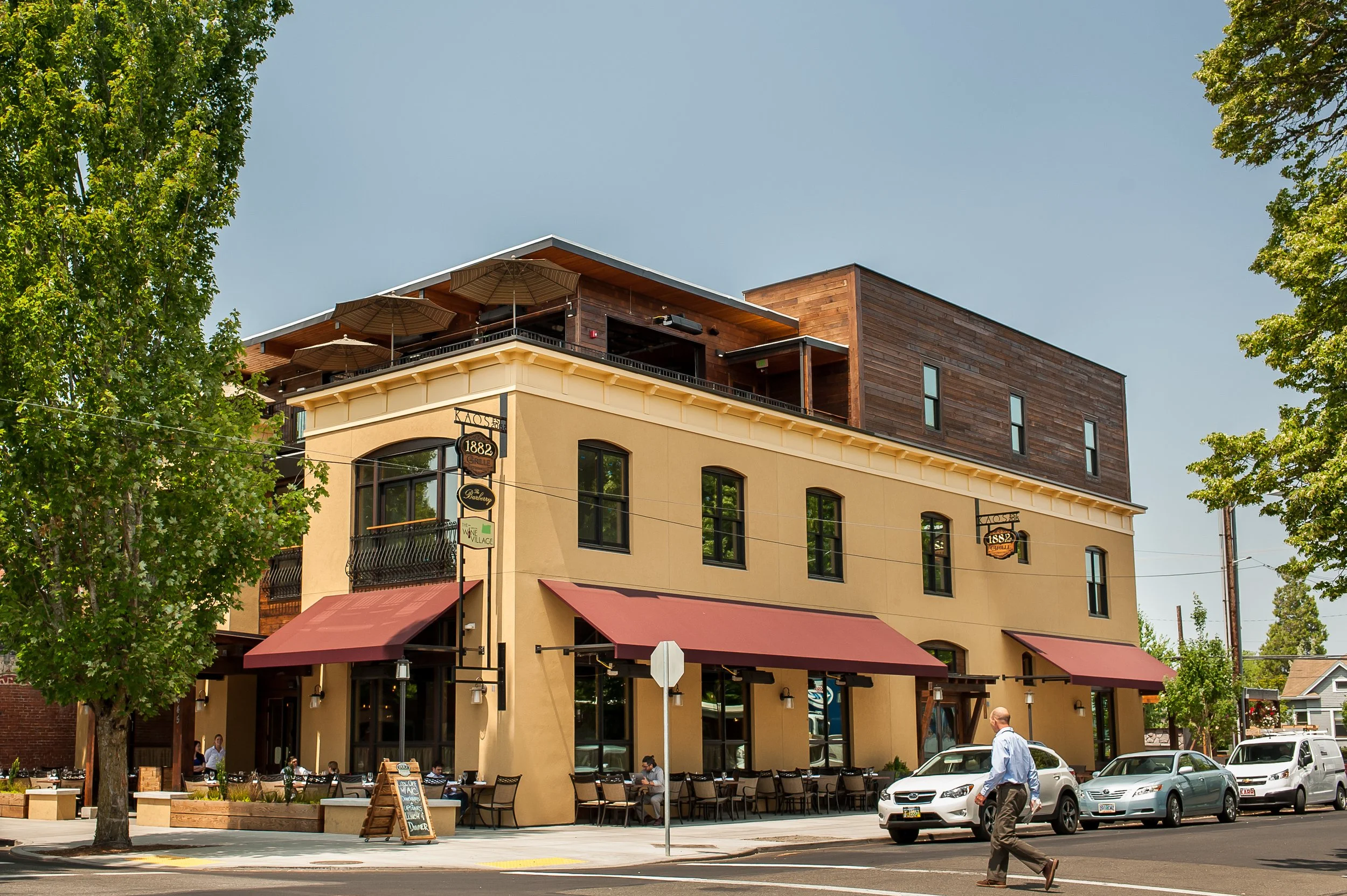 A three-story building with yellow and brown exterior, awnings, and outdoor seating, located on a main street with pedestrians and parked cars.