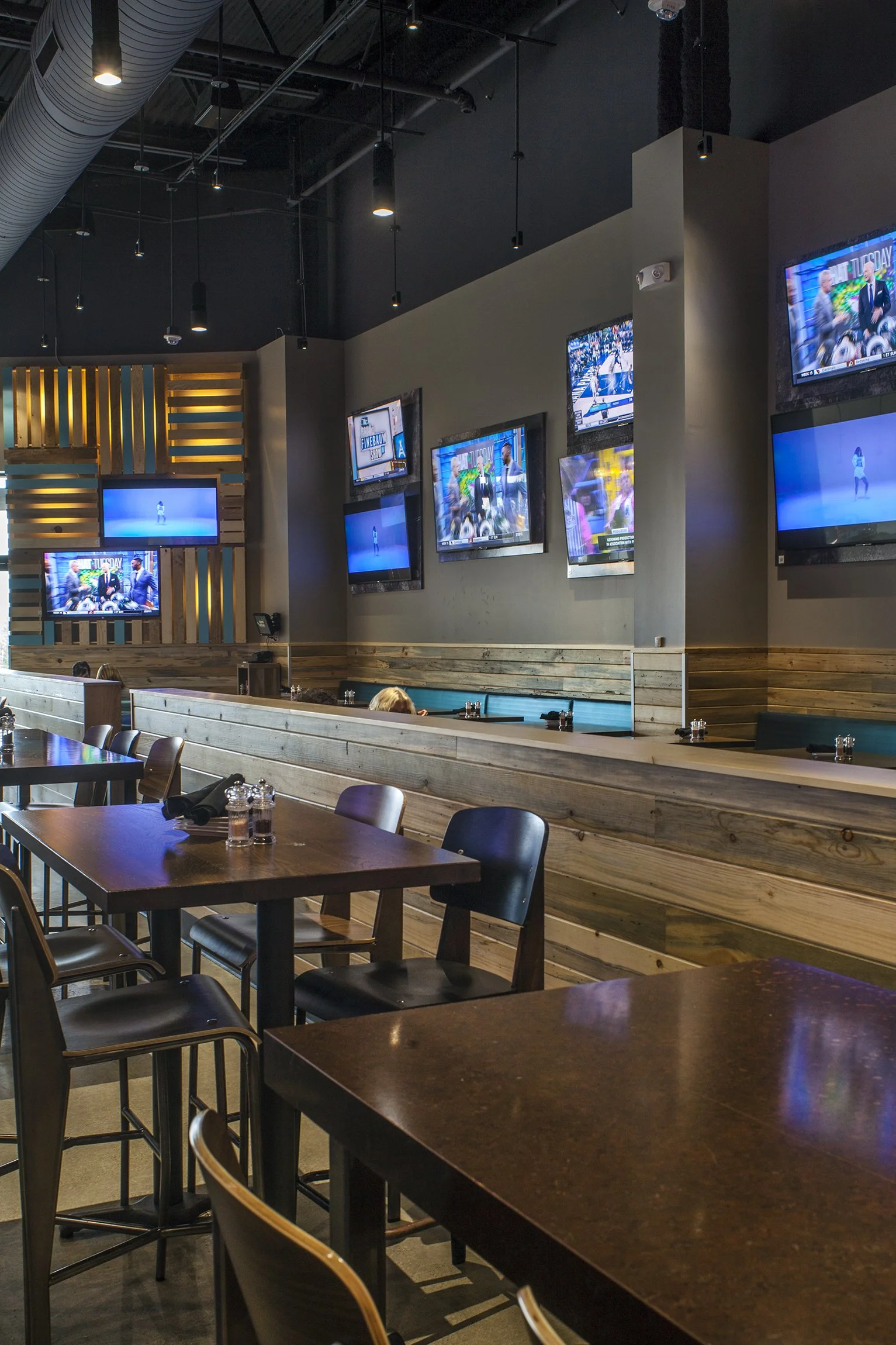 Inside a modern bar or restaurant with multiple TVs on the wall showing sports or entertainment content, wooden tables and chairs, and dim lighting.