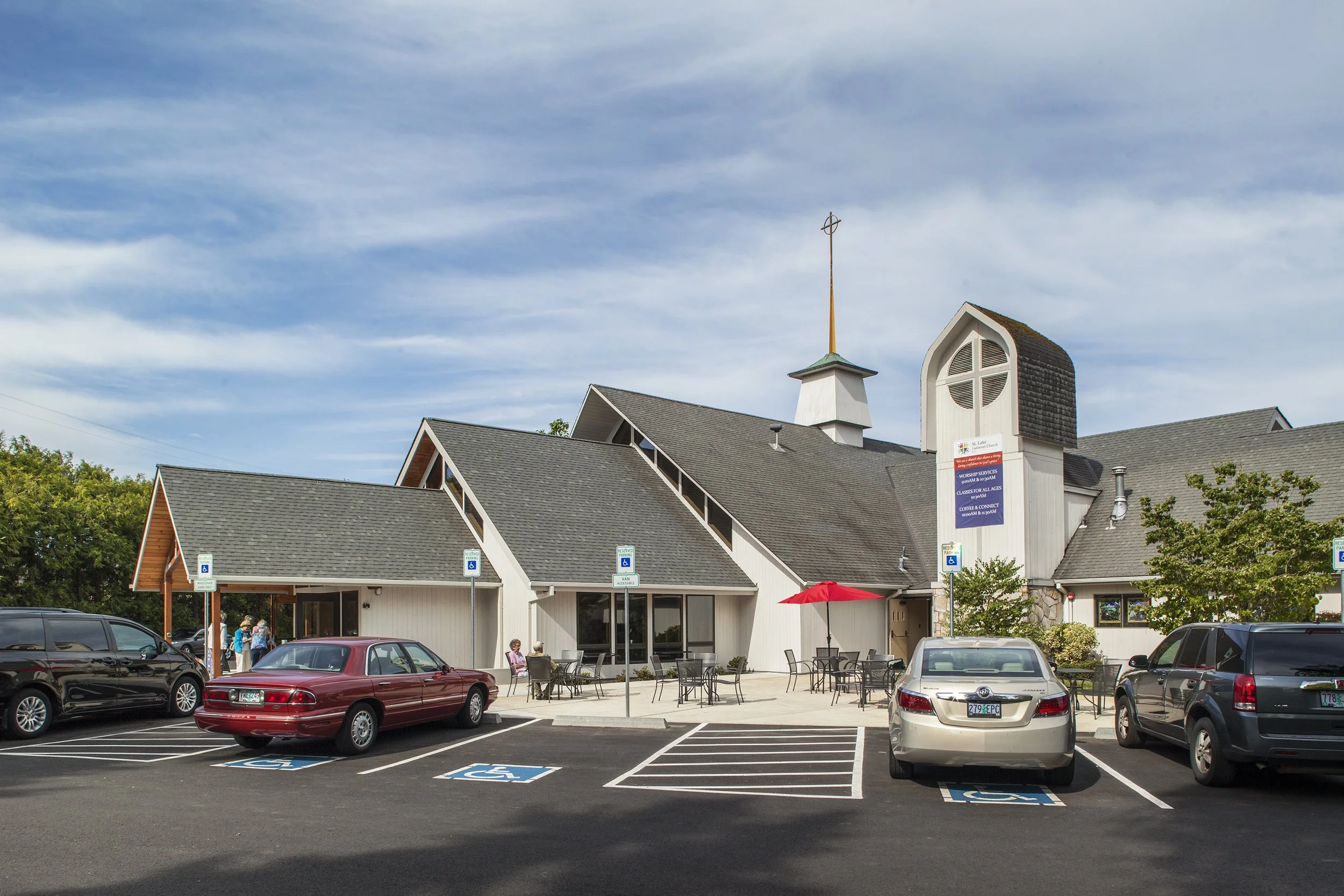 Church building with a parking lot in front, several handicap-accessible parking spaces, and a few people sitting outside at tables on a sunny day.
