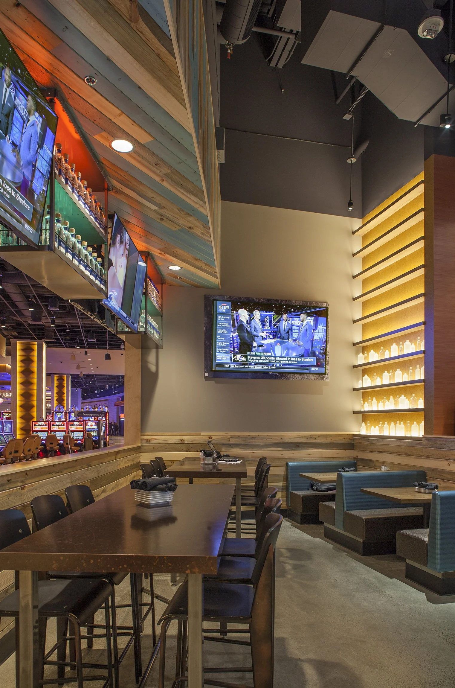 Interior of a restaurant or bar with a large wooden table, chairs, booth seating, and multiple flat-screen TVs mounted on the walls showing sports and news.