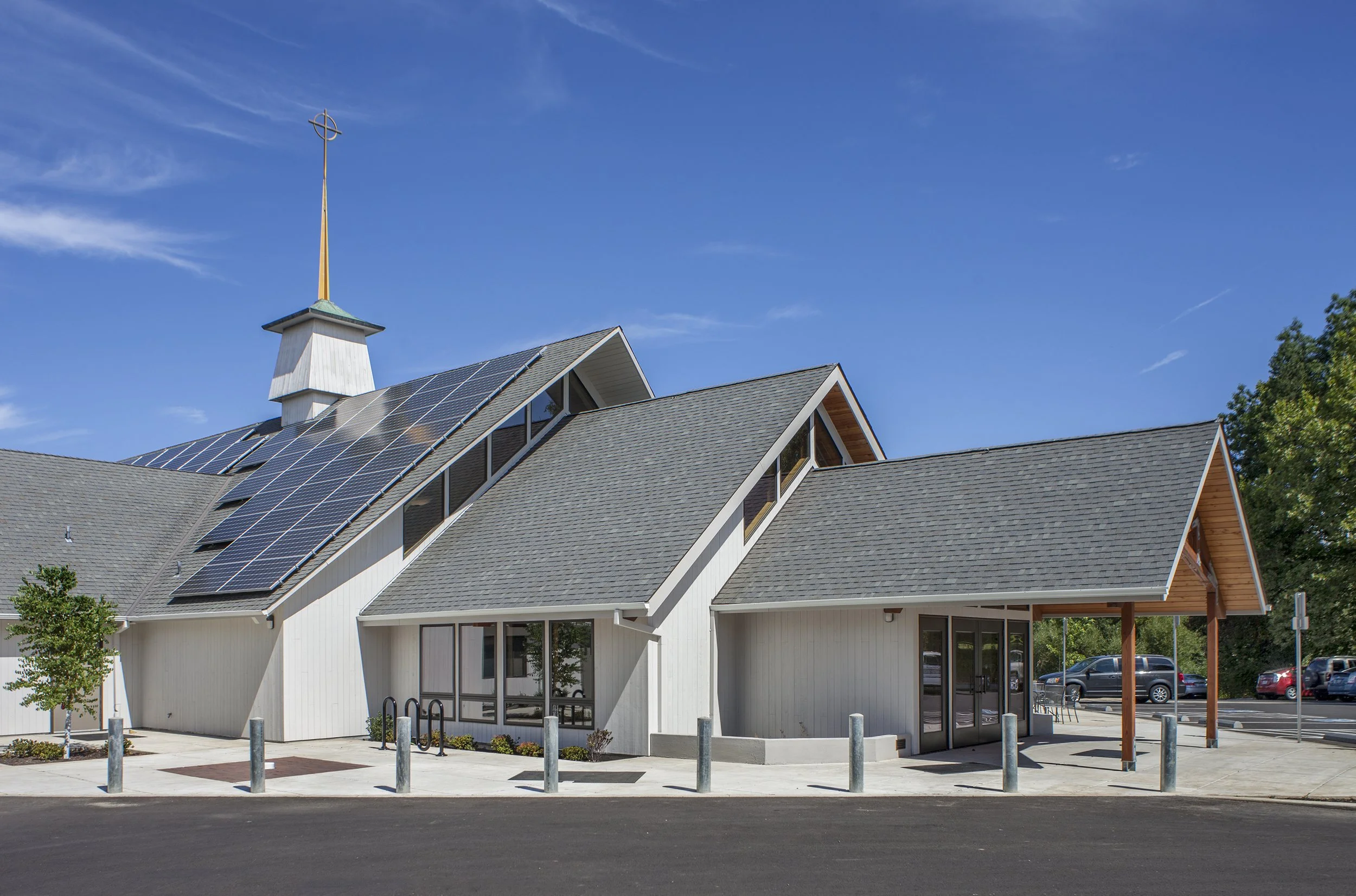 Modern church building with solar panels, gray roof, white exterior, glass entrance doors, and a small parking lot under a clear blue sky.