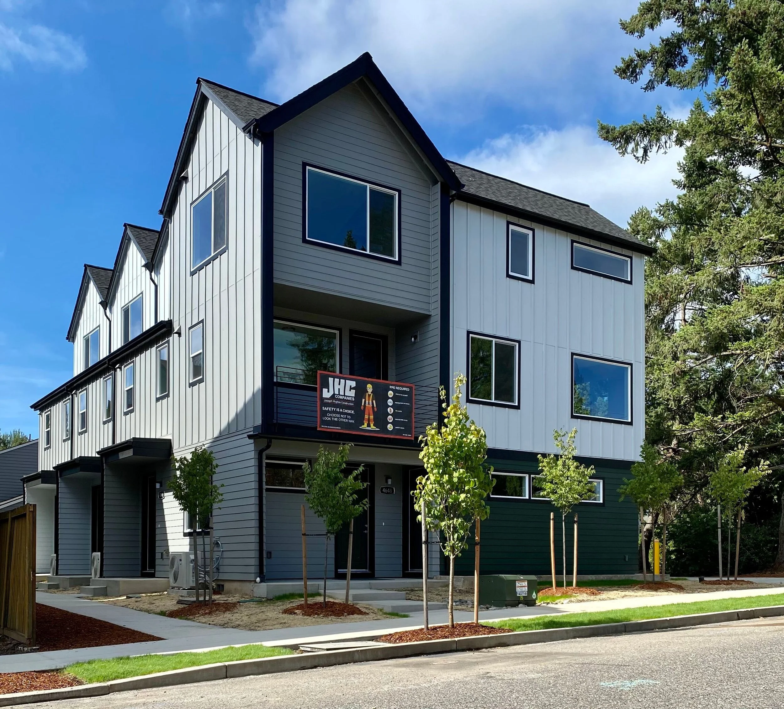 New multi-story residential building with modern design, white and dark green exterior, multiple windows, small front yard with young trees, sidewalk, and street in foreground.
