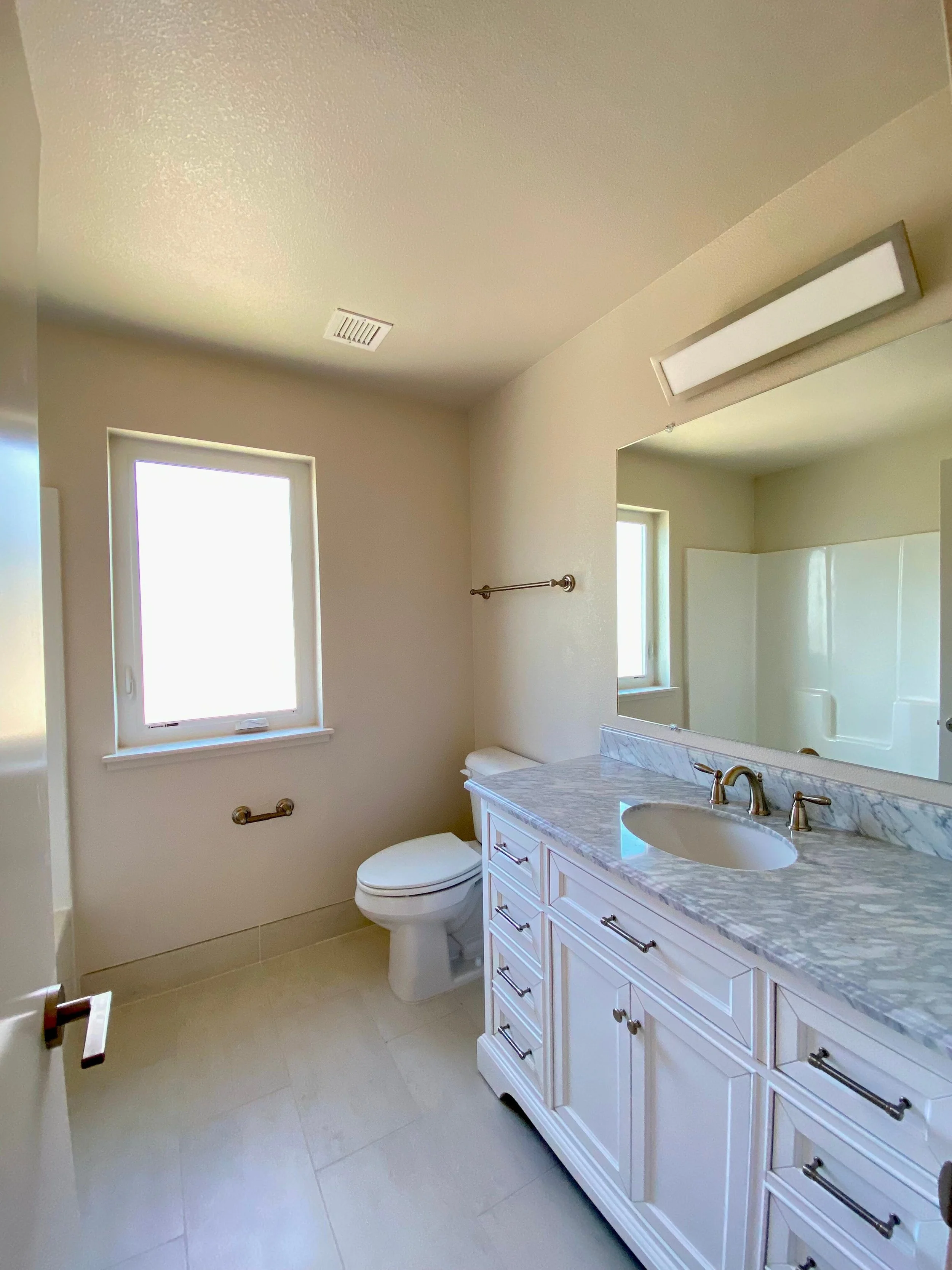 Bright bathroom with a window, white toilet, white vanity with a marble countertop, large mirror, and a shower area with a glass door.