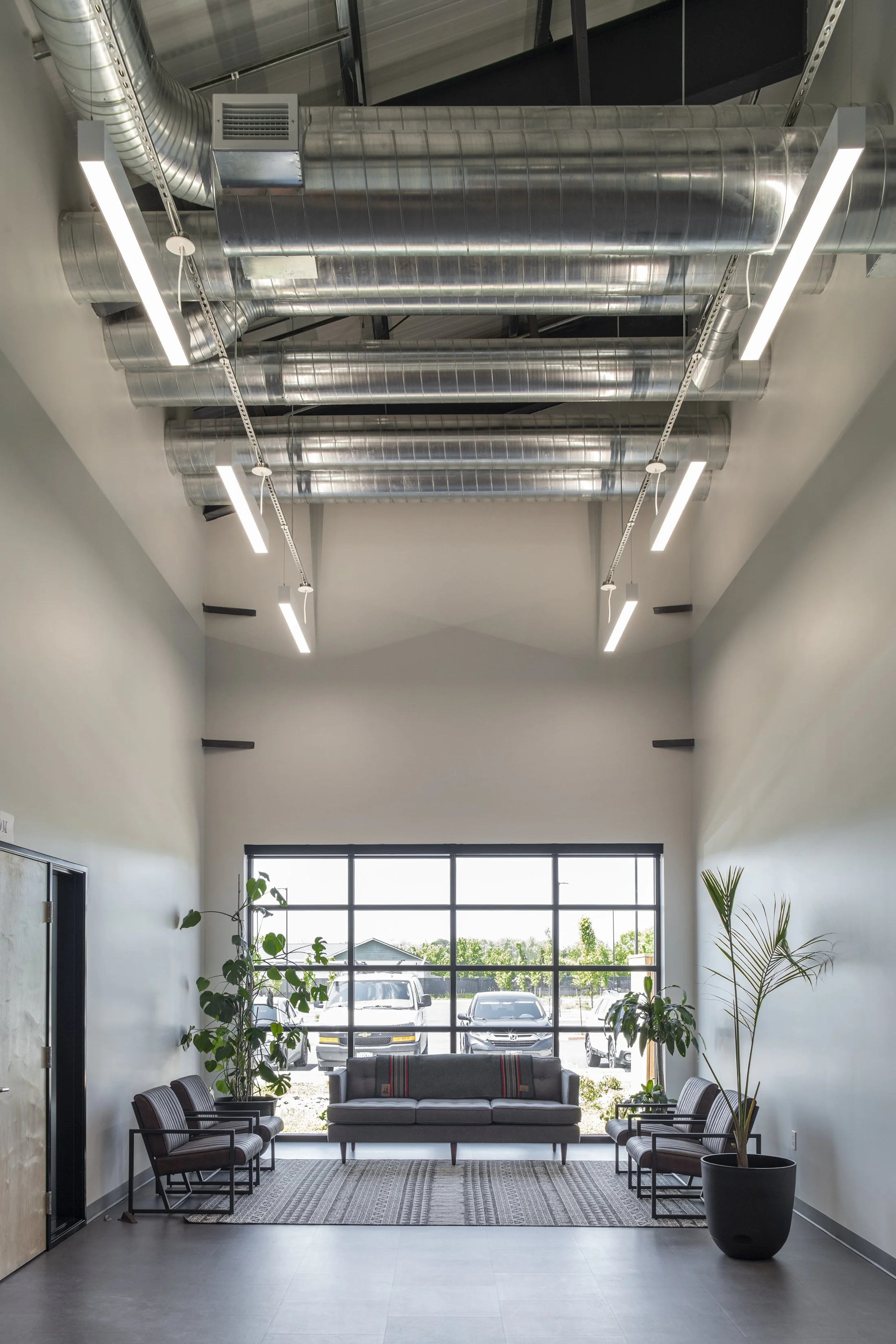 Modern waiting room with large front window, gray sofa, four chairs, potted plants, and exposed ductwork ceiling.